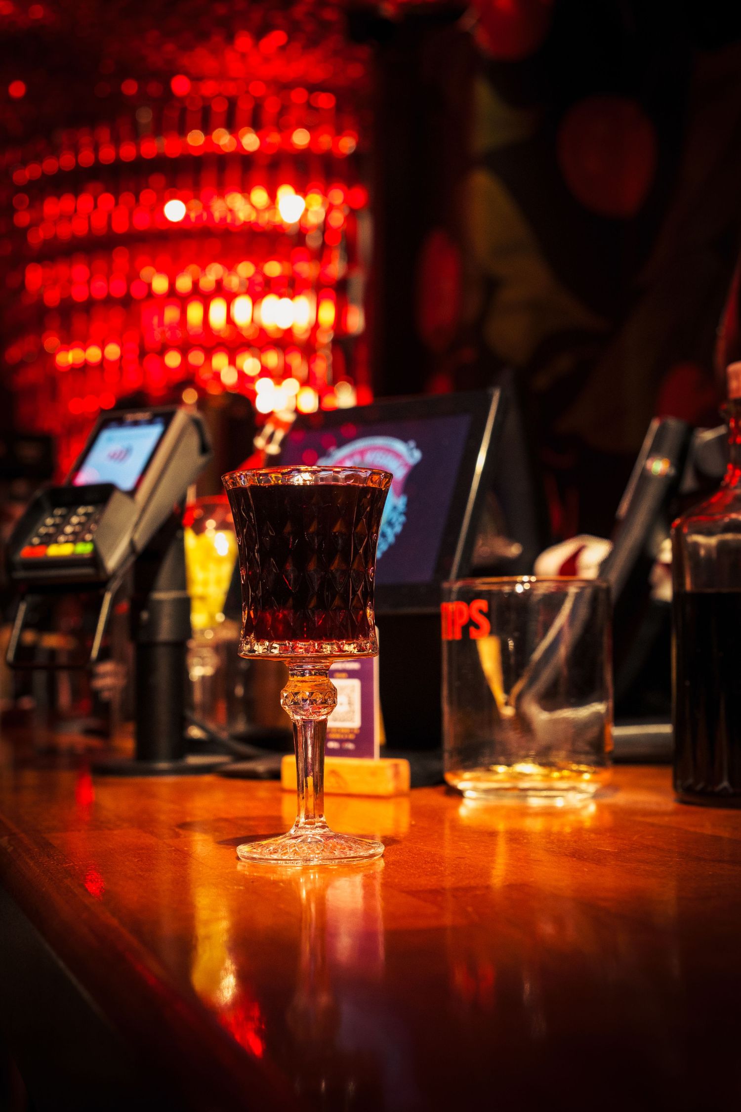 A dark cocktail in an ornate glass sits on a polished bar counter with red ambient lighting and payment terminals in background.