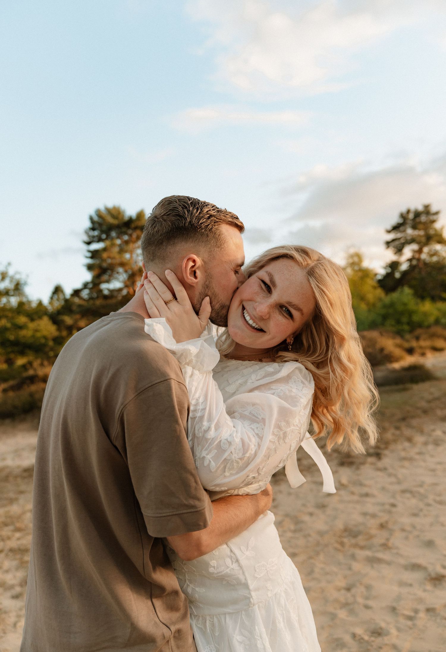 Een romantisch stel tijdens een fotoshoot op het strand bij zonsondergang. De vrouw draagt een witte jurk en straalt terwijl ze wordt gekust