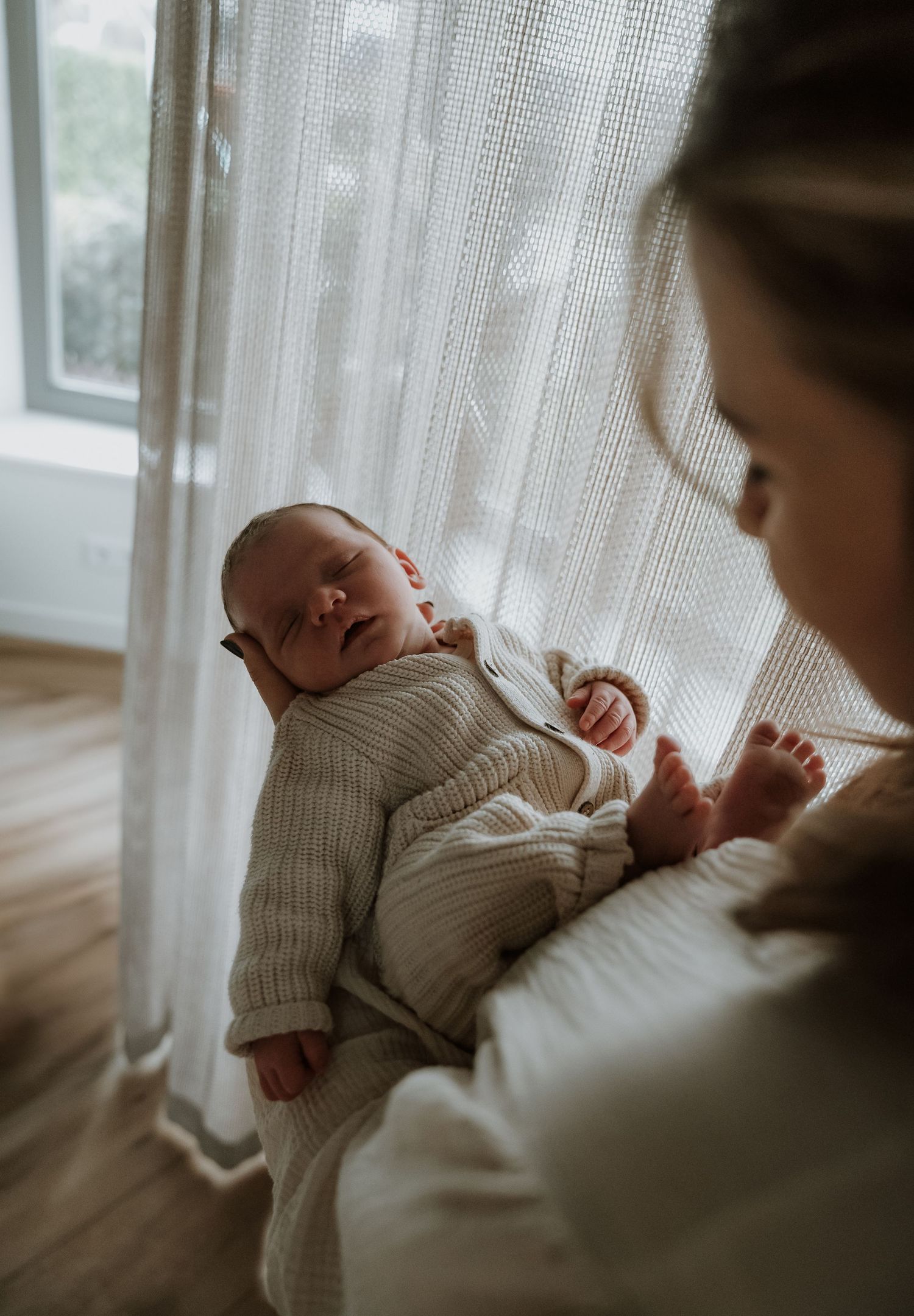 Moeder houdt slapende pasgeboren baby vast bij raam met gordijn, gefotografeerd door Myrthegwenfotografie.