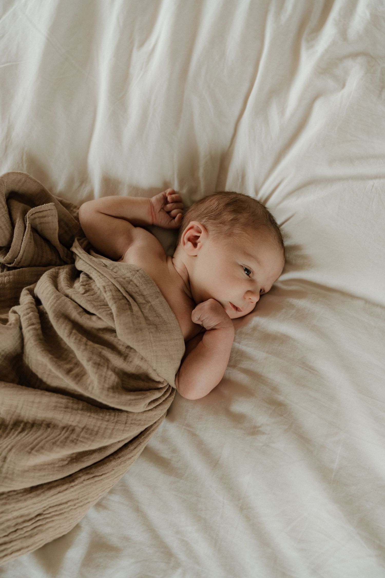 Rustgevend portret van een wakkere pasgeboren baby, ingewikkeld in een zachte beige doek op een licht bed, vastgelegd in een pure newborn.