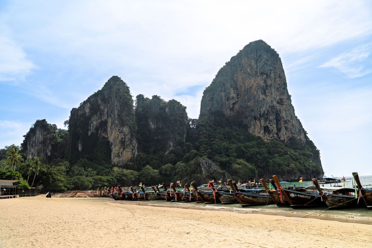 Towering limestone cliffs and long-tail boats at Railay Beach in Thailand with tourists on the shore.