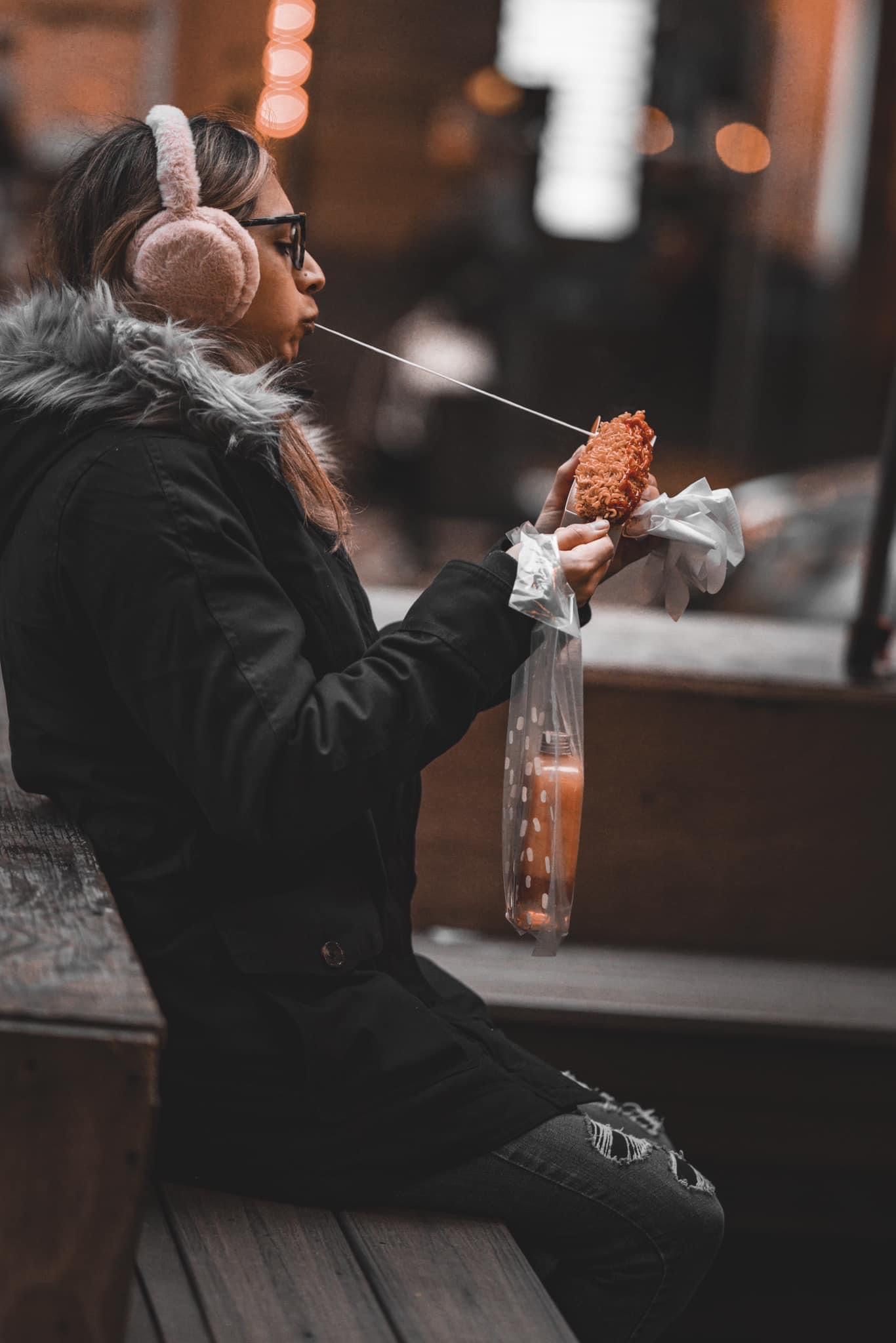 Person in black jacket enjoying food at outdoor seating area with bokeh lights.