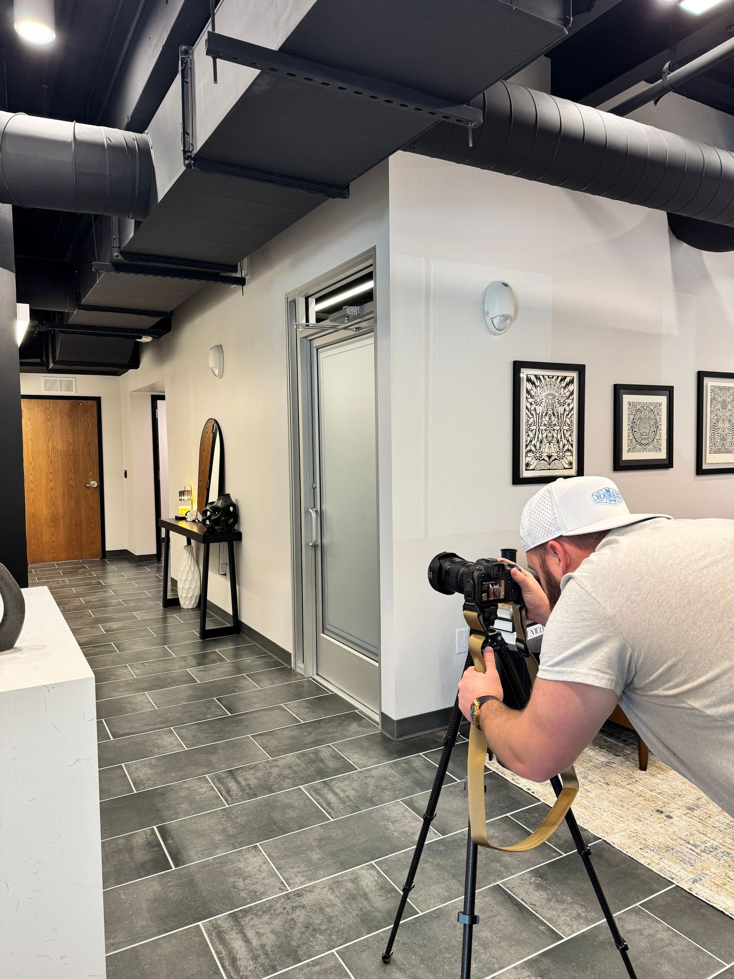 Professional photographer crouching to capture low-angle shots in a modern hallway with dark tile floors and white walls.