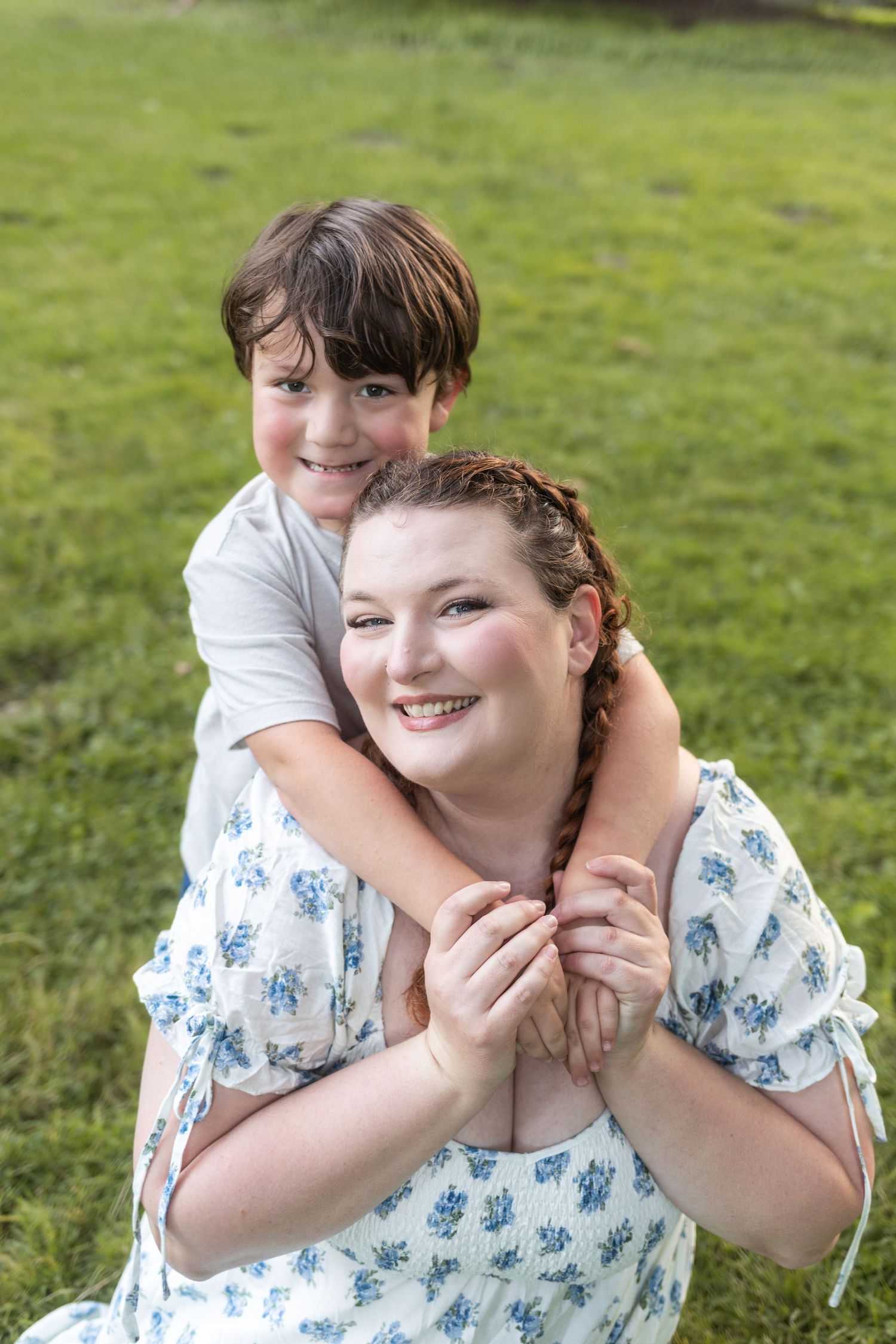 A young boy hugs a smiling woman in a floral dress from behind while sitting on green grass outdoors.