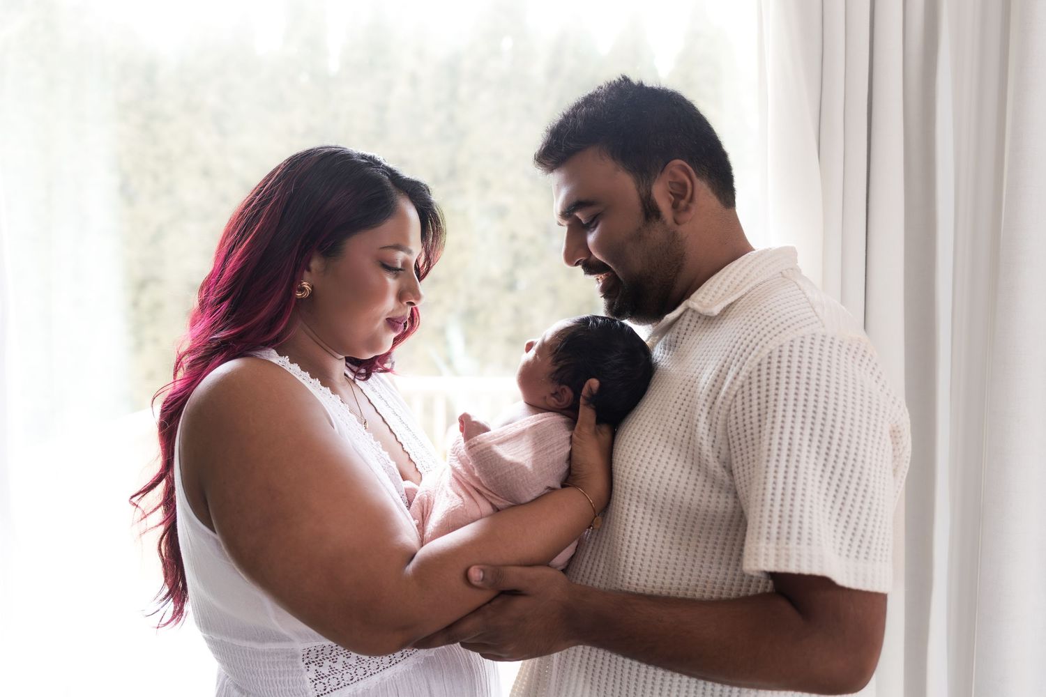 A couple tenderly holds their newborn baby near a bright window in a soft, intimate family portrait.