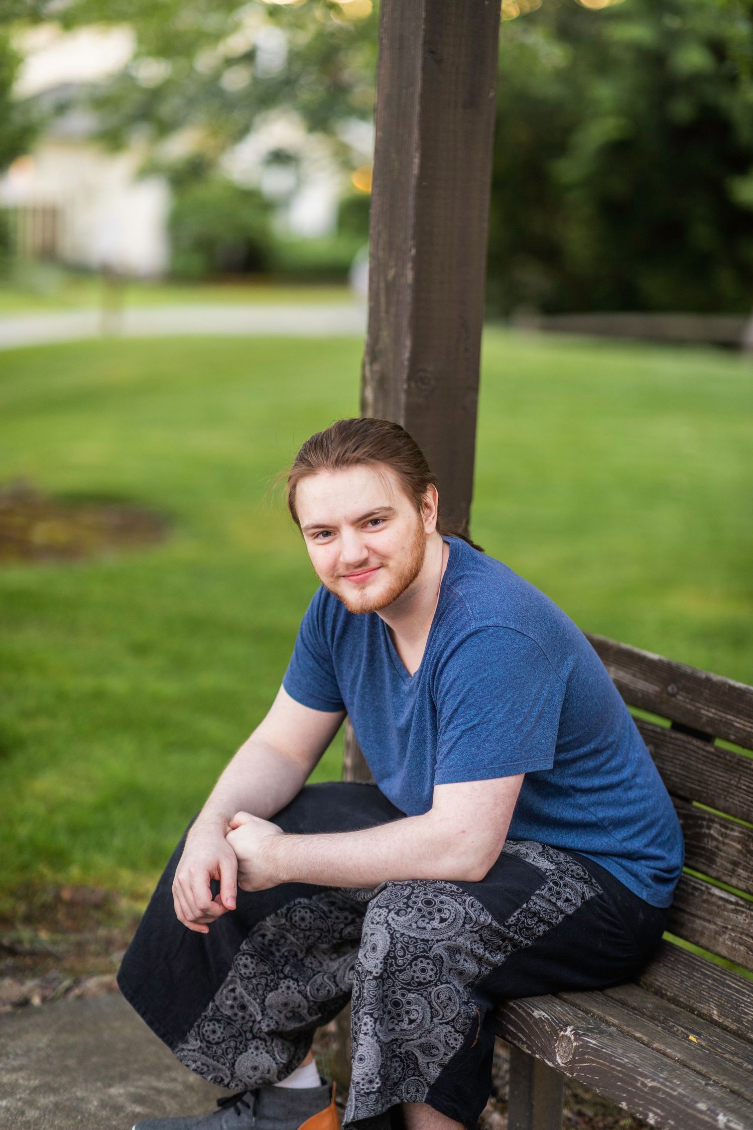 Young man wearing a blue shirt stands and smiles in front of a blurry green background.