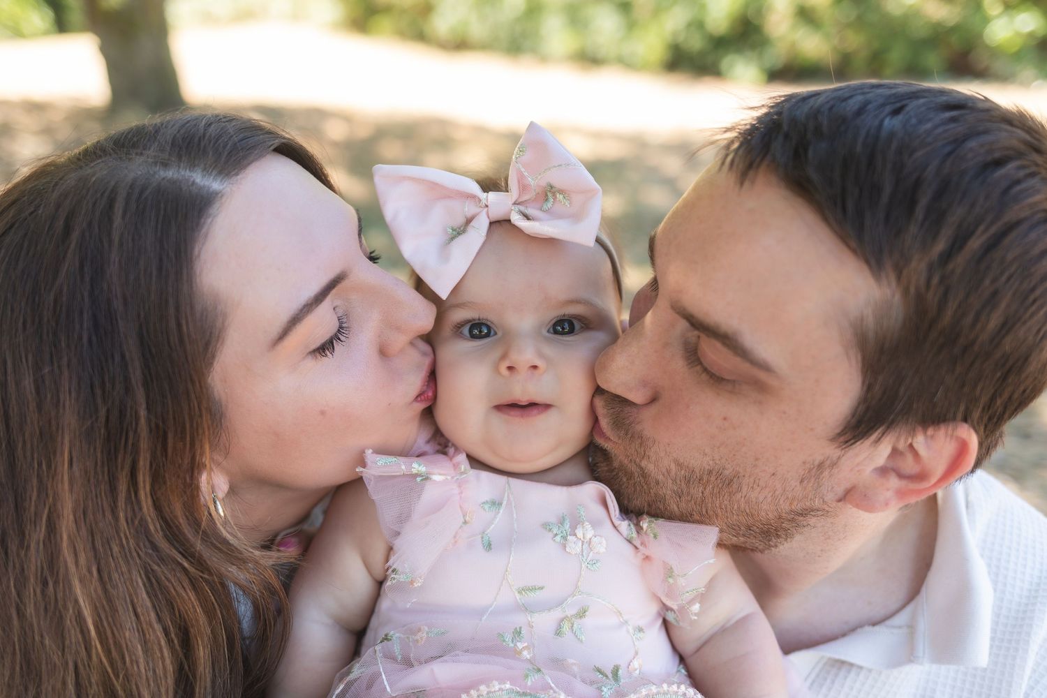 Parents kissing their adorable baby girl wearing a pink bow and floral dress in an outdoor park setting.