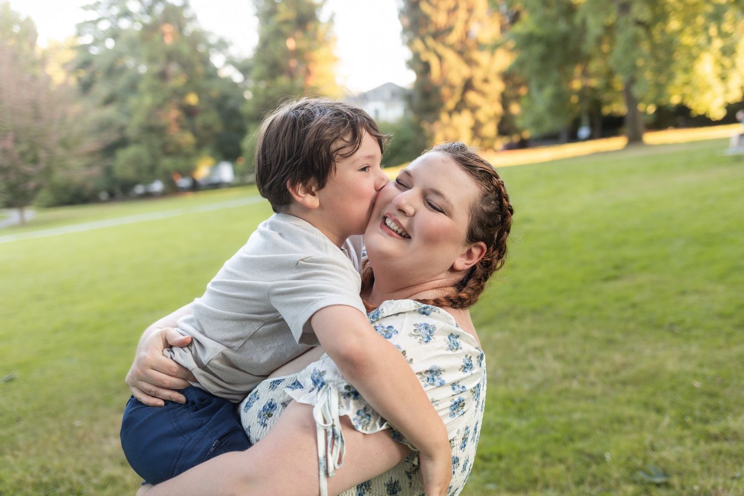 A young boy joyfully kisses his smiling mother on the cheek while she holds him in a sunny green park.