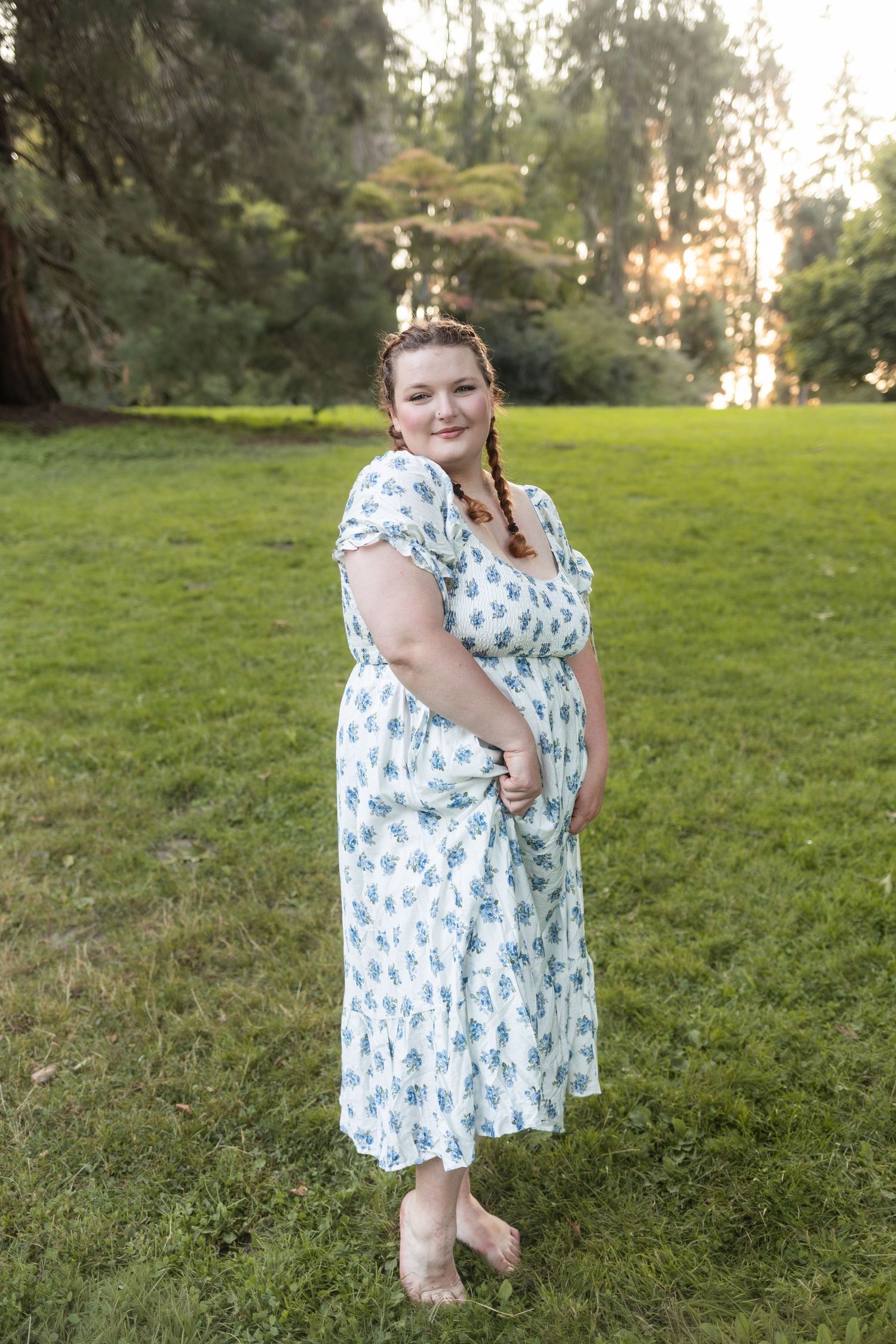 Woman in floral midi dress standing barefoot on green grass in a sunlit park with tall trees.