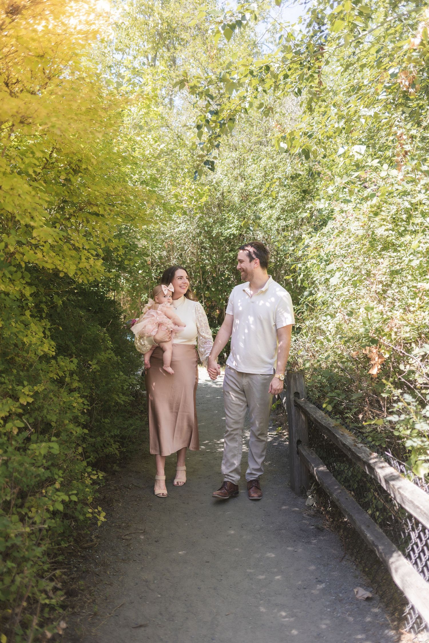A couple holds hands and walks along a garden path, with the woman carrying a baby in a pink outfit on a sunny day.