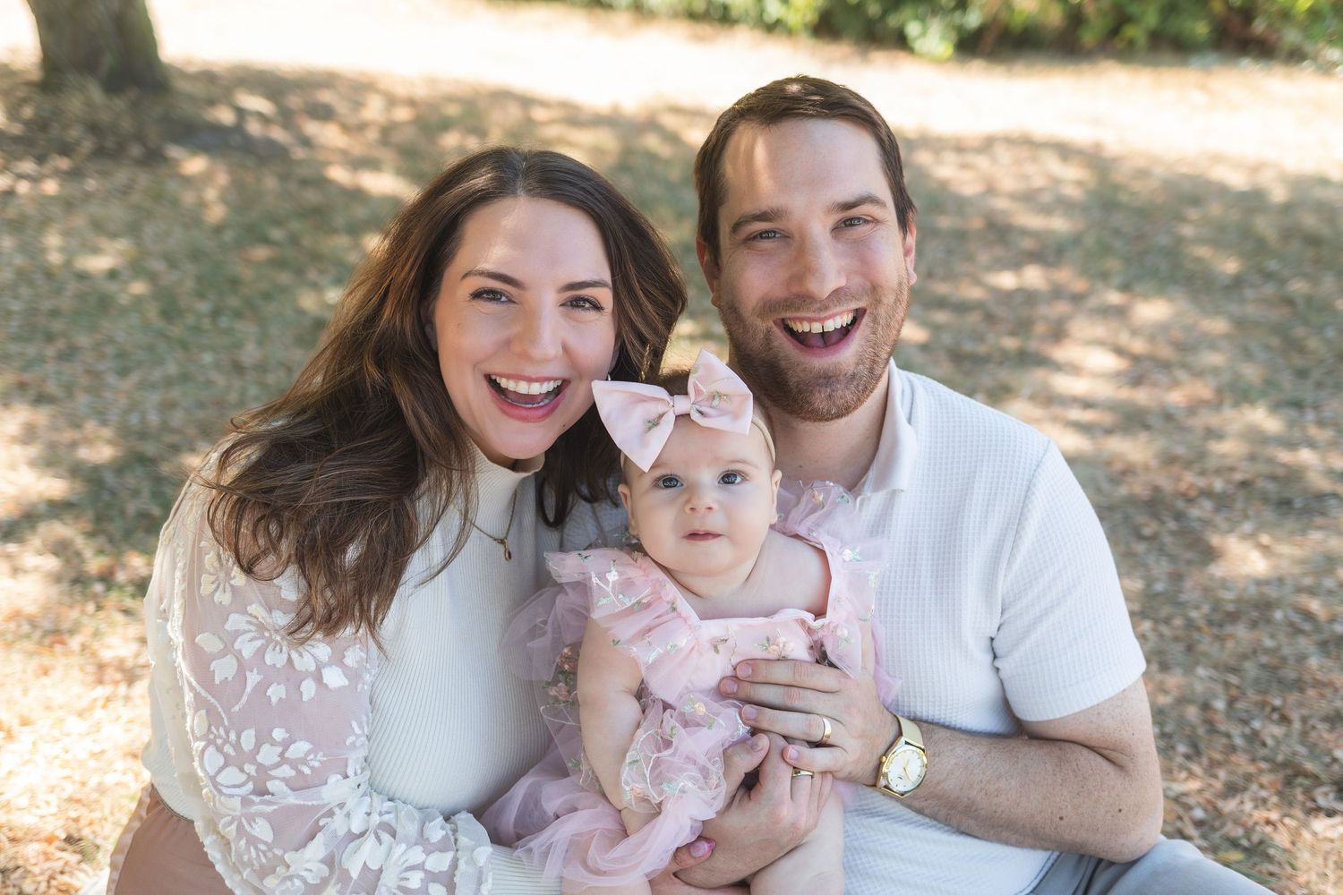 Happy young family with laughing parents holding their baby girl in a pink dress outdoors in a park.
