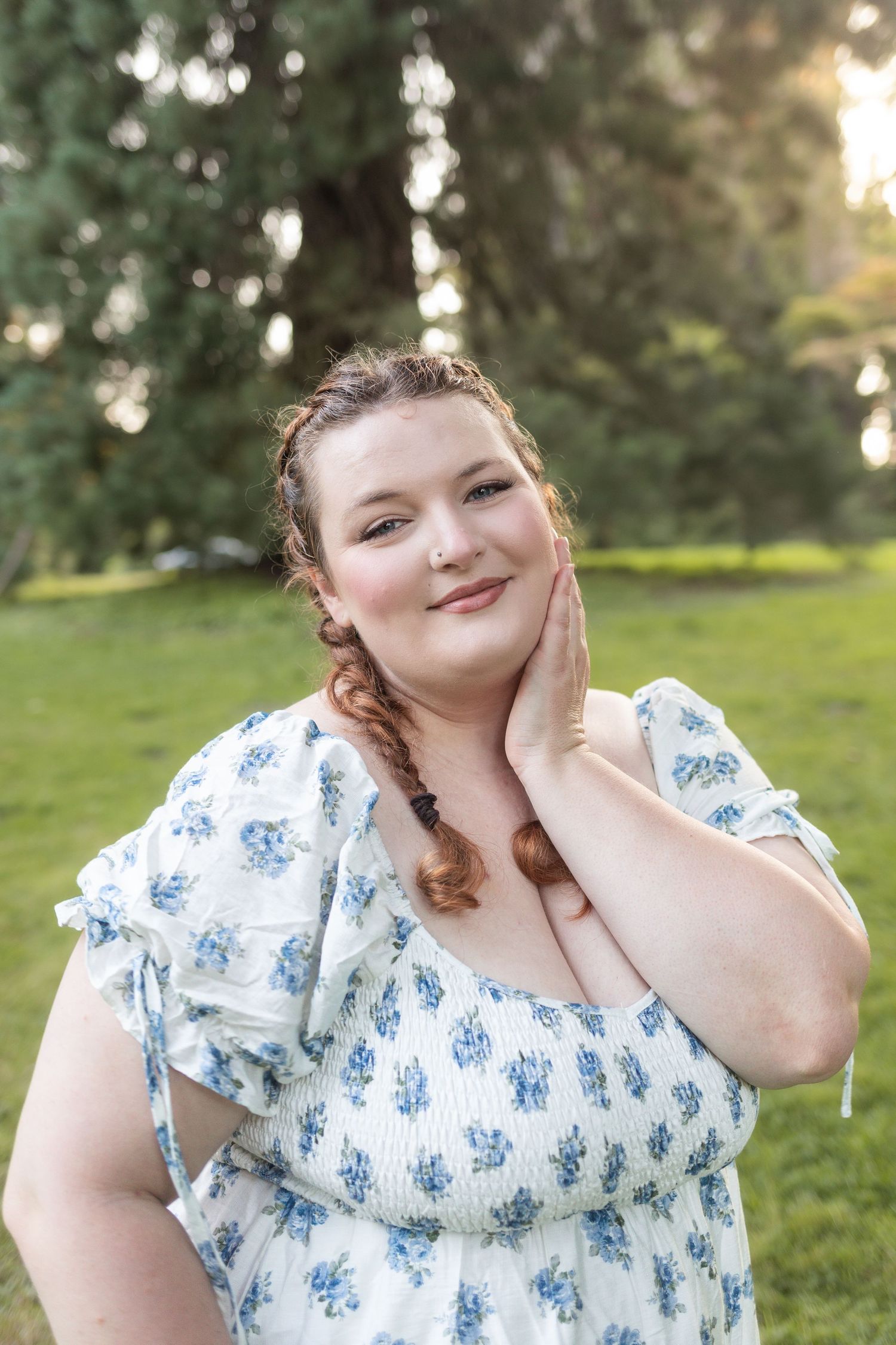 A plus-size woman in a white floral dress smiles warmly in a sunlit park, touching her neck with braided red hair.
