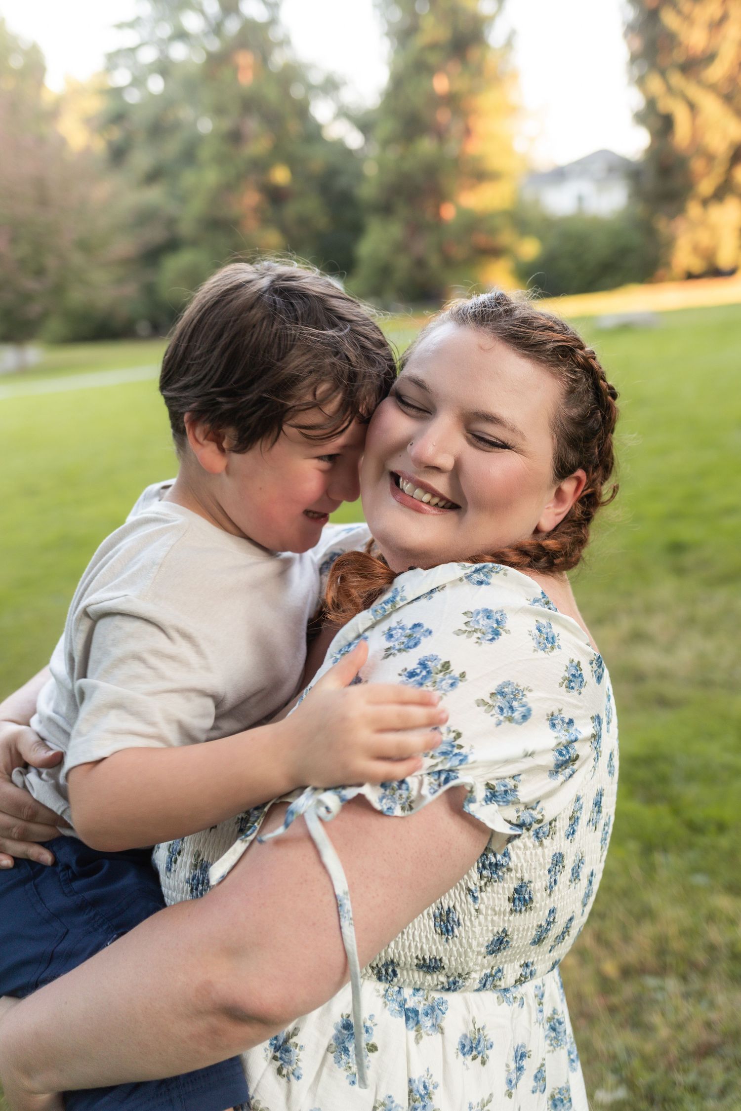 A smiling woman in a floral dress joyfully hugs a young boy outdoors in a sunny green park.
