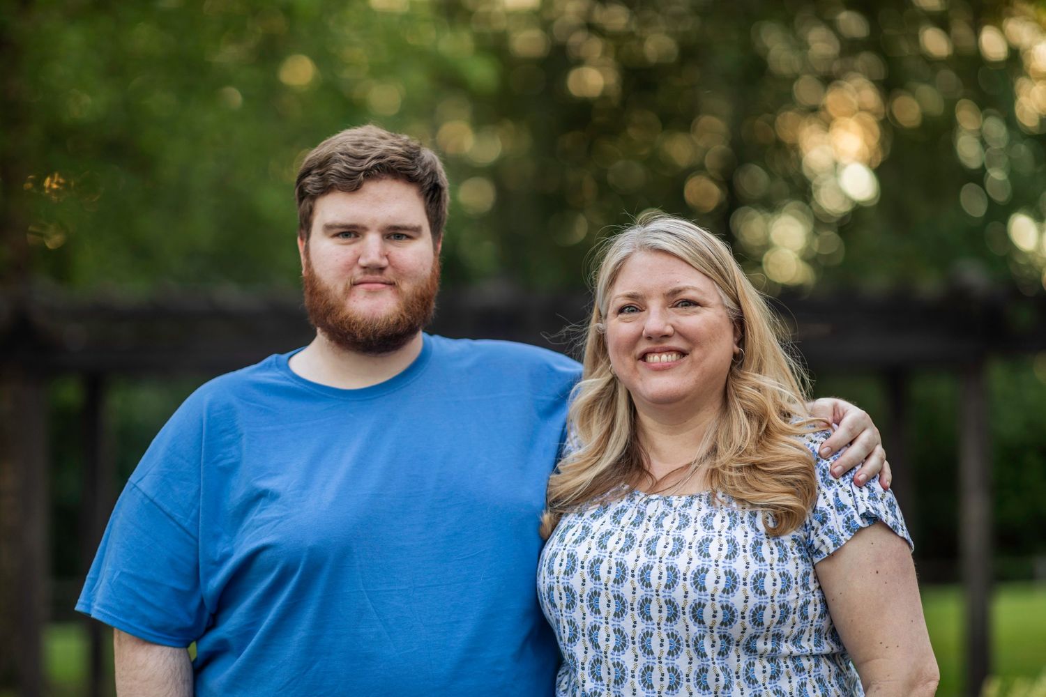 Bushy bearded young man wearing a blue shirt stands and smiles in front of a blurry green background with his blonde haired mother.