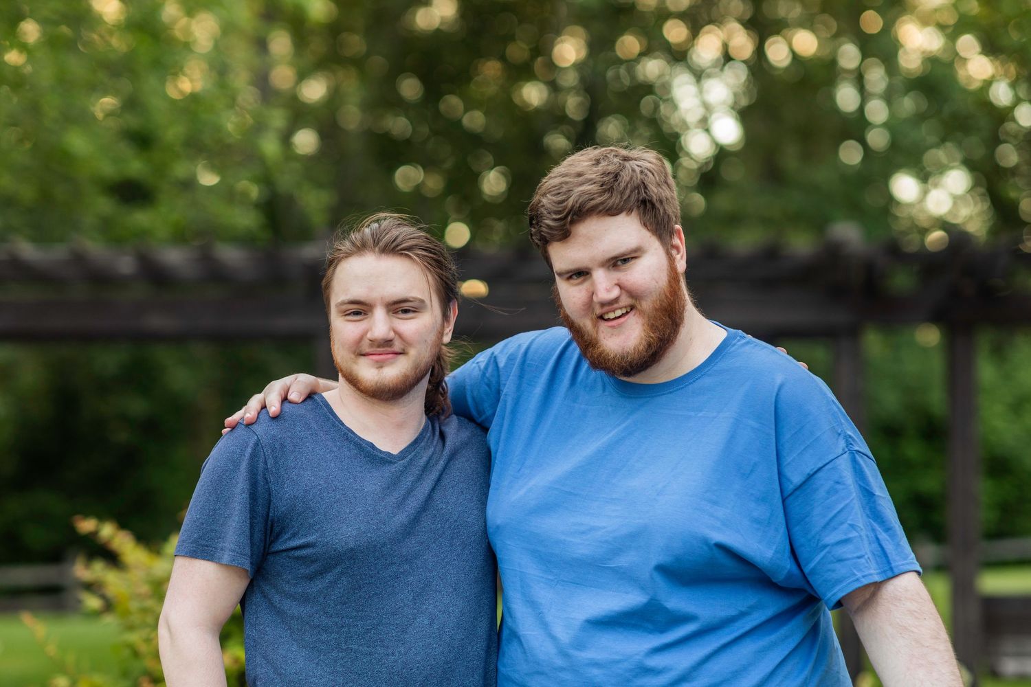 Bushy bearded young man wearing a blue shirt stands and smiles in front of a blurry green background with his little brother.