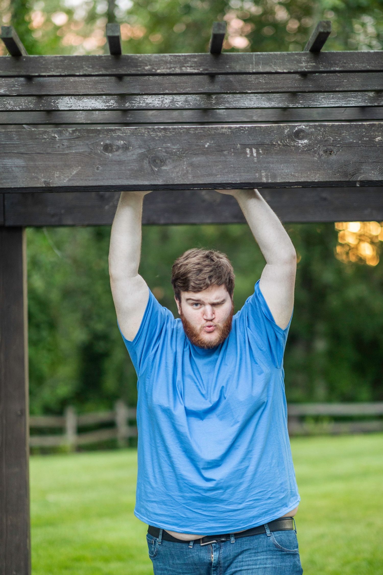 Bushy bearded young man wearing a blue shirt stands and smiles in front of a blurry green background.