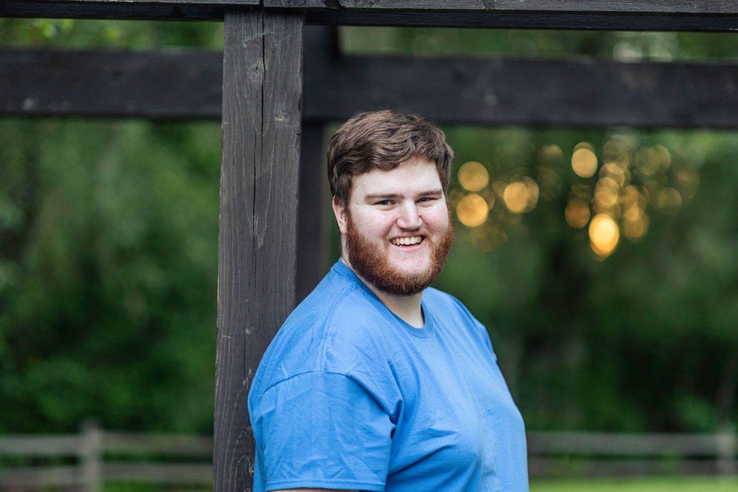 Bushy bearded young man wearing a blue shirt stands and smiles in front of a blurry green background.