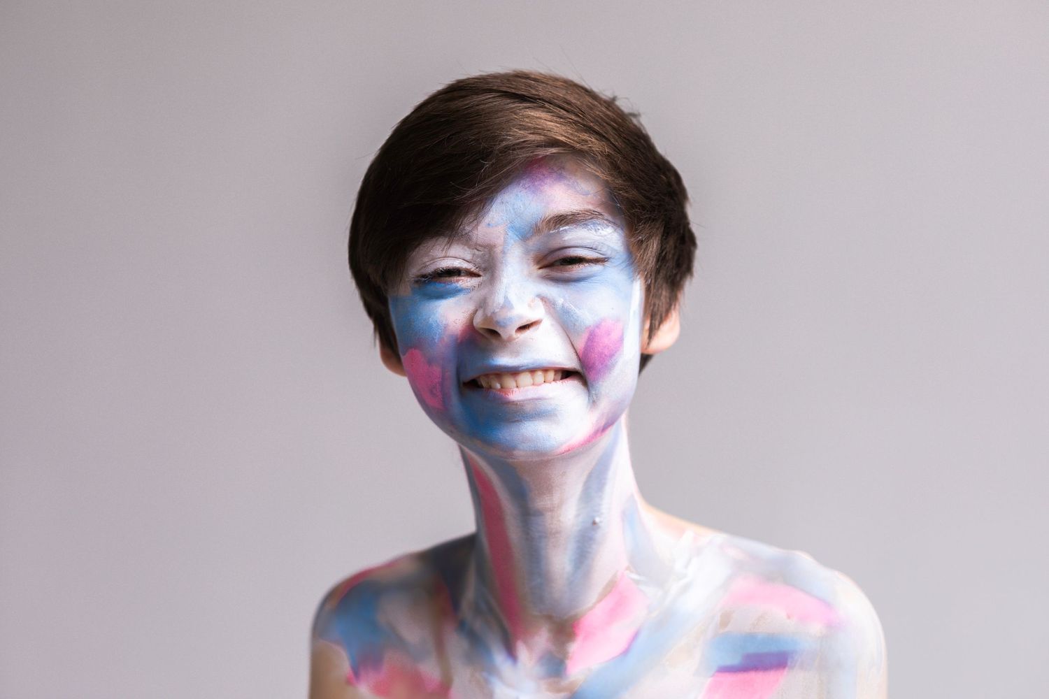 Short-haired individual is painted with the colors of the transgender flag and smiles widely.