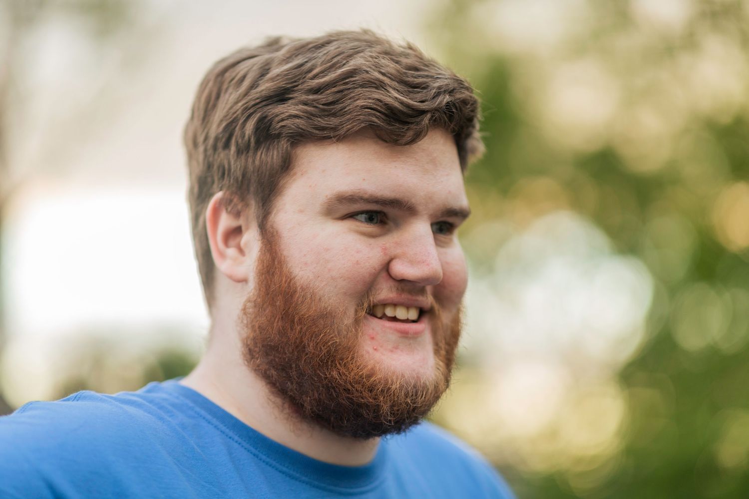Bushy bearded young man wearing a blue shirt stands and smiles in front of a blurry green background.