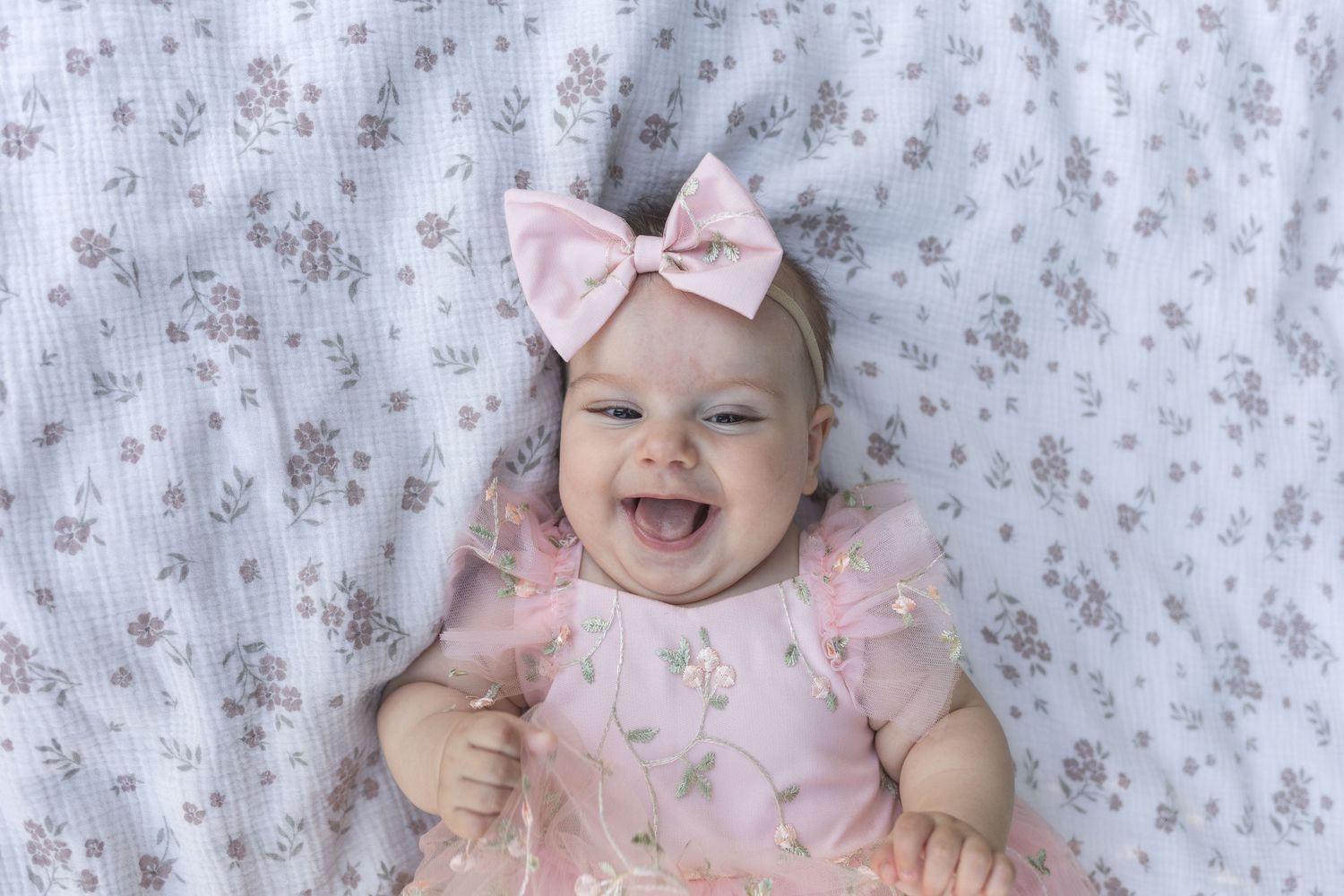 Smiling baby girl in pink dress and bow headband lying on floral blanket.
