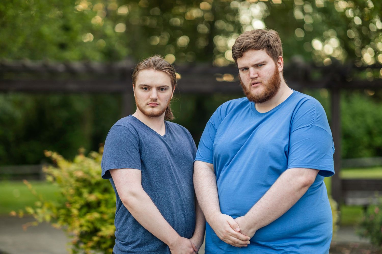 Bushy bearded young man wearing a blue shirt stands and poses seriously in front of a blurry green background with his brother.