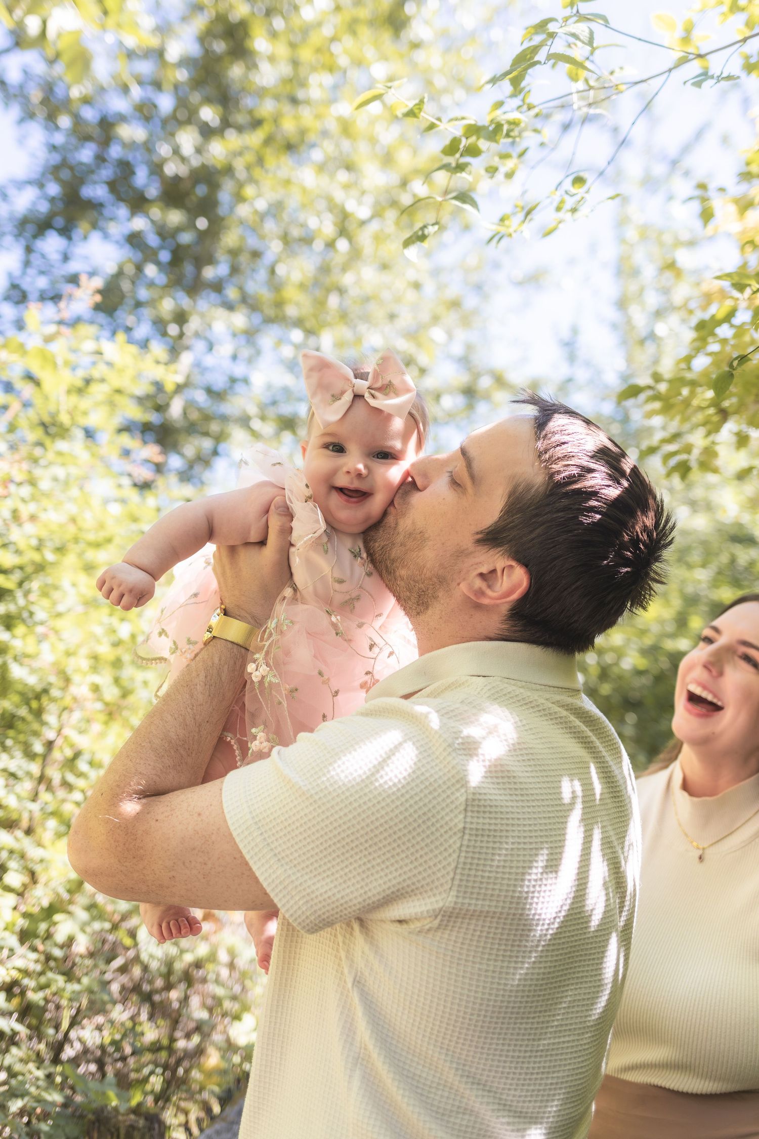 A father kisses his smiling baby girl in a pink dress outdoors, while a woman laughs beside them.