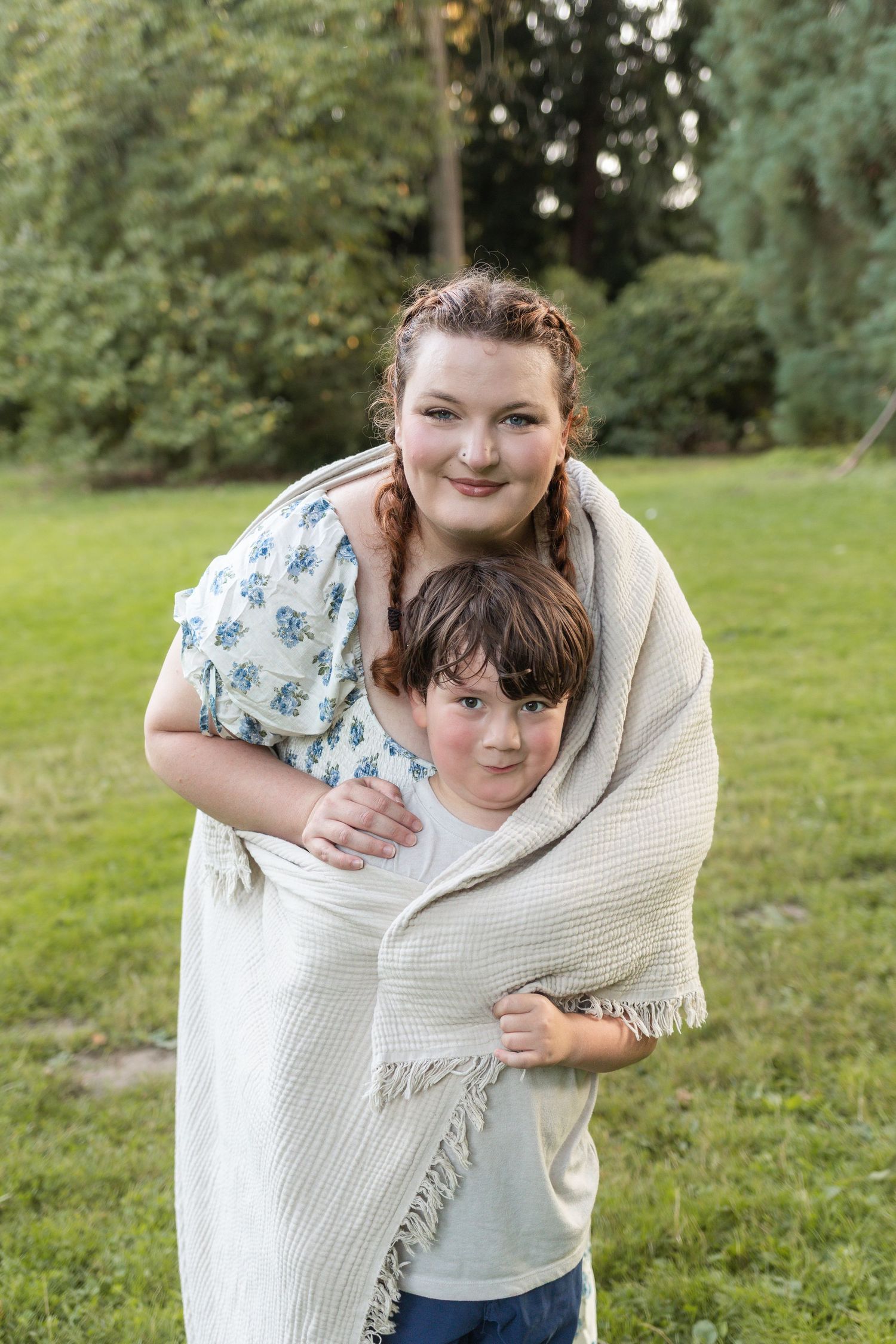 Teen girl and young boy wrapped together in a cozy blanket smiling outdoors in a lush green park.