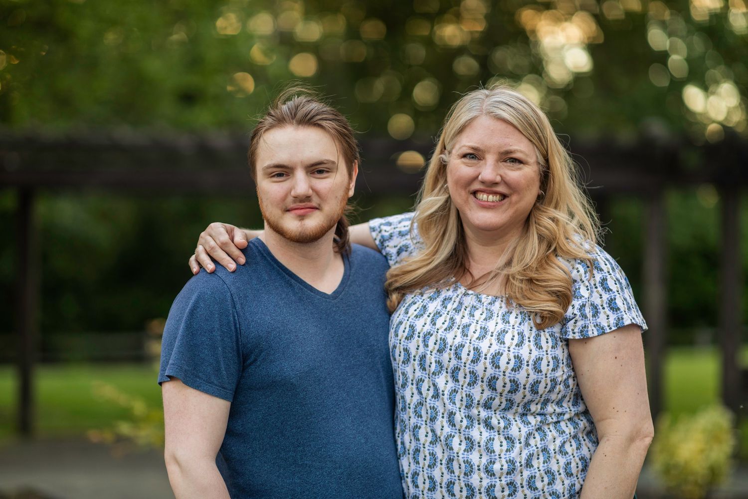 Young man wearing a blue shirt stands and smiles in front of a blurry green background with his mother.
