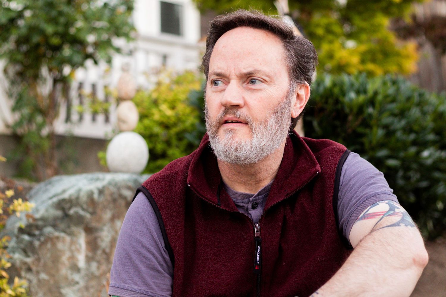 50-something man with a gray beard looks off into the distance with stacked rocks behind him.