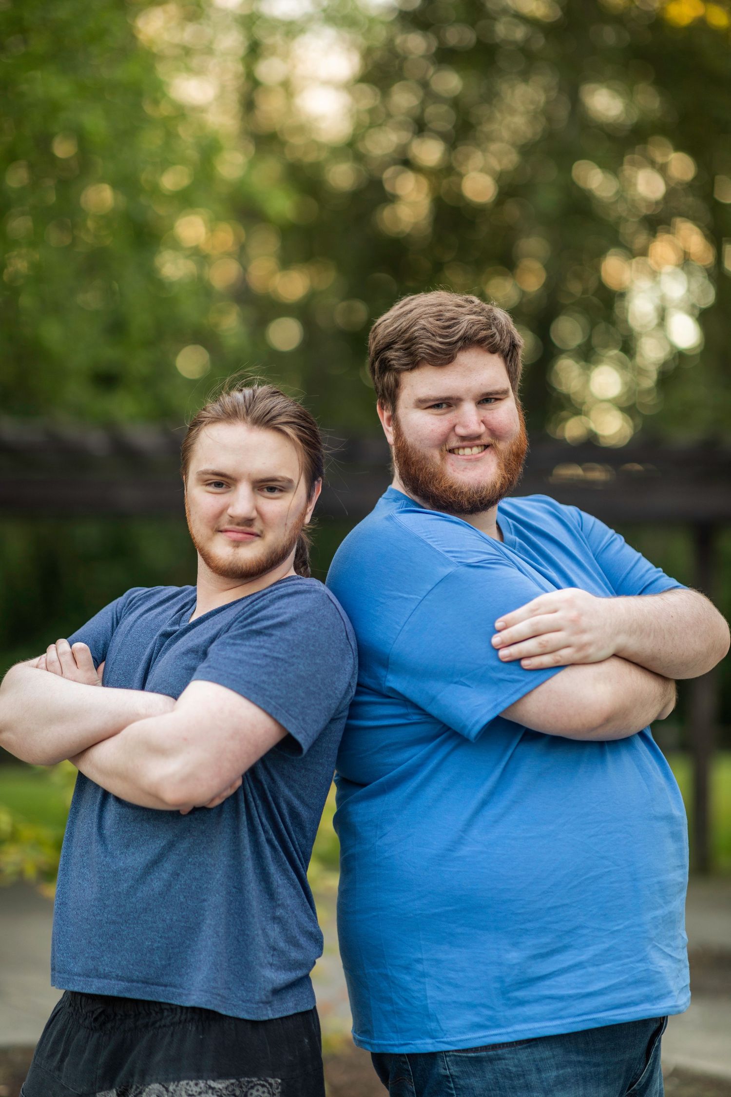 Bushy bearded young man wearing a blue shirt stands and smiles in front of a blurry green background with his little brother.