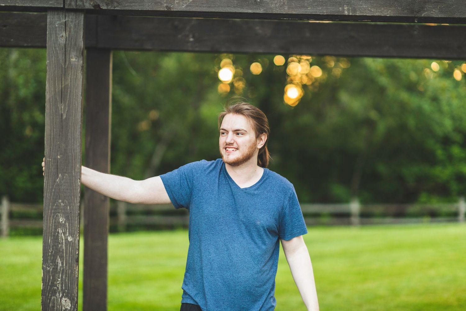 Non-binary individual smiles and looks off to the distance with blurry green grass and trees in the background.