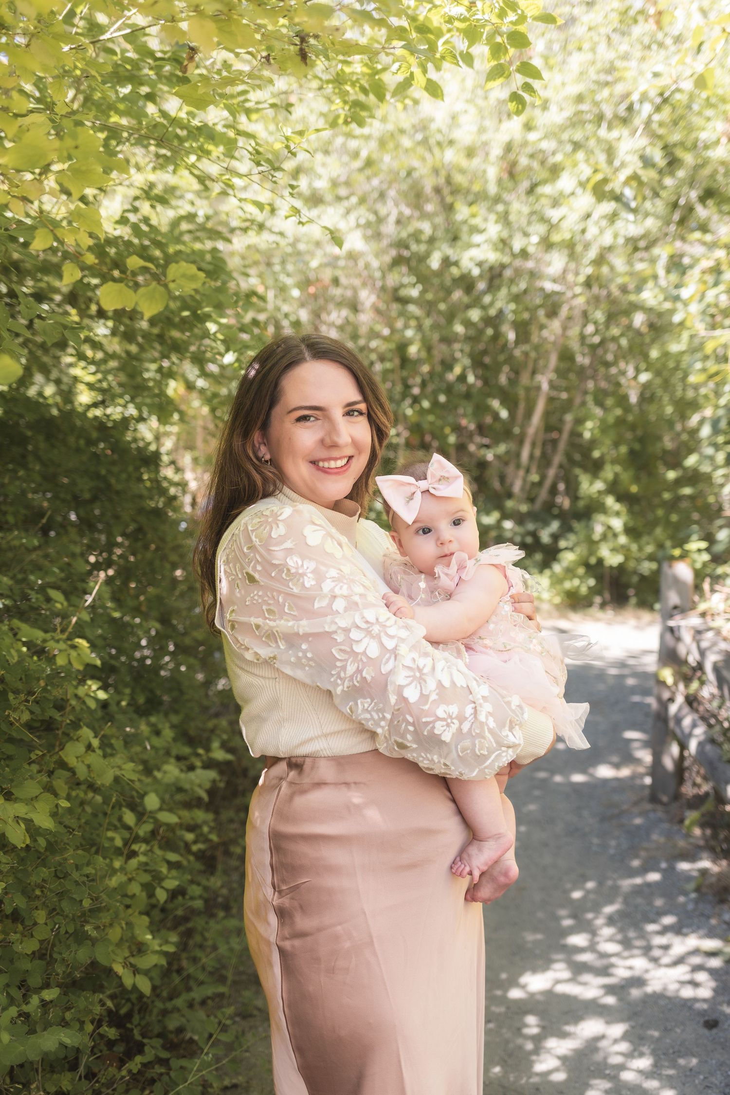 A smiling woman in a floral lace top holds a baby girl in a pink dress outdoors among lush green trees.