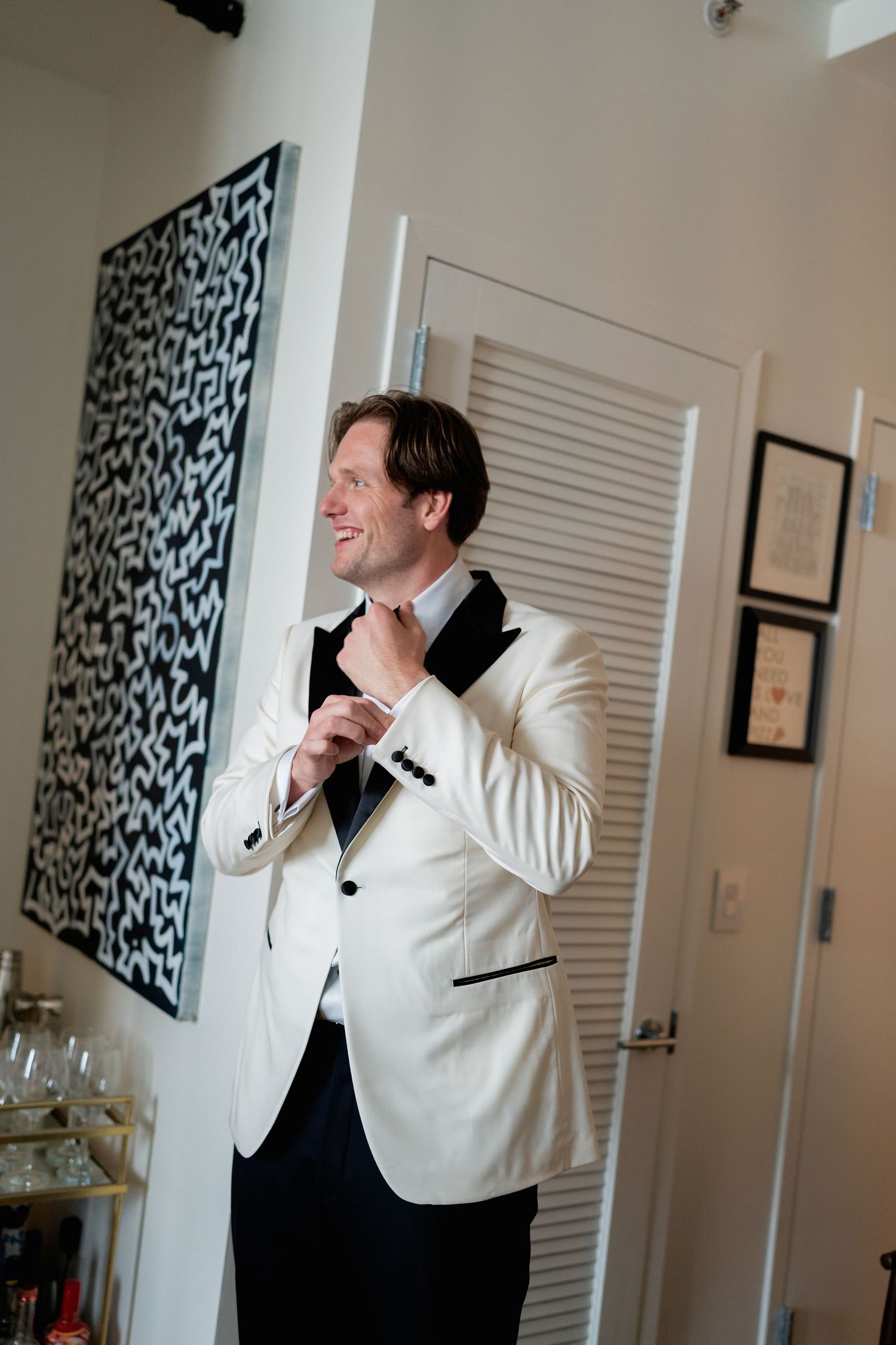 A person in a white tuxedo jacket adjusts their bowtie while getting ready in a room with modern art on the wall.