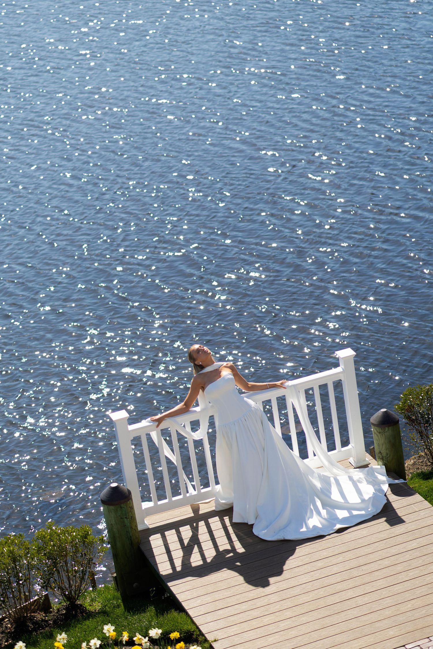 Bride in white gown standing on dock overlooking sparkling water.