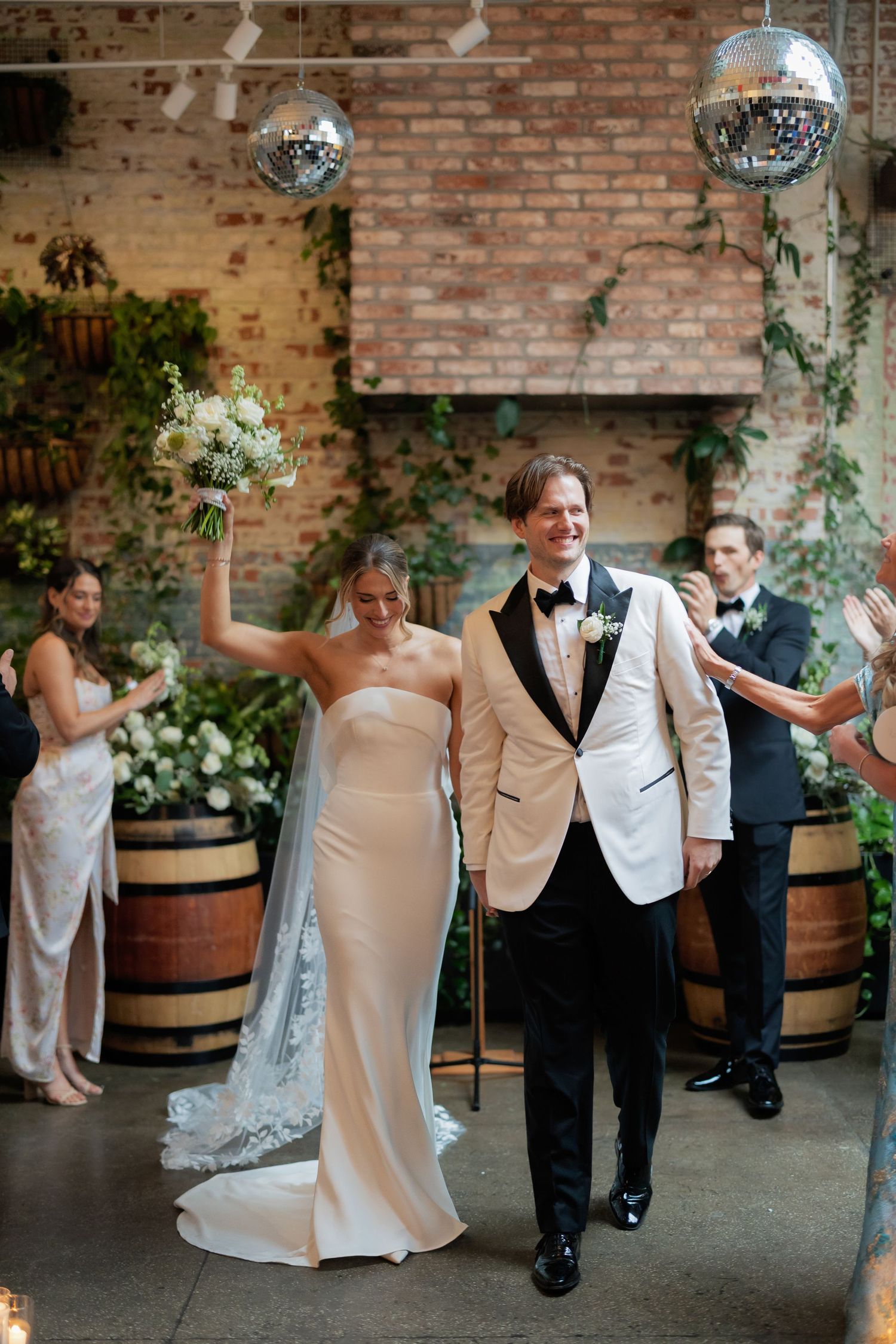 Wedding couple dances joyfully in a rustic brick-walled venue decorated with disco balls and greenery.