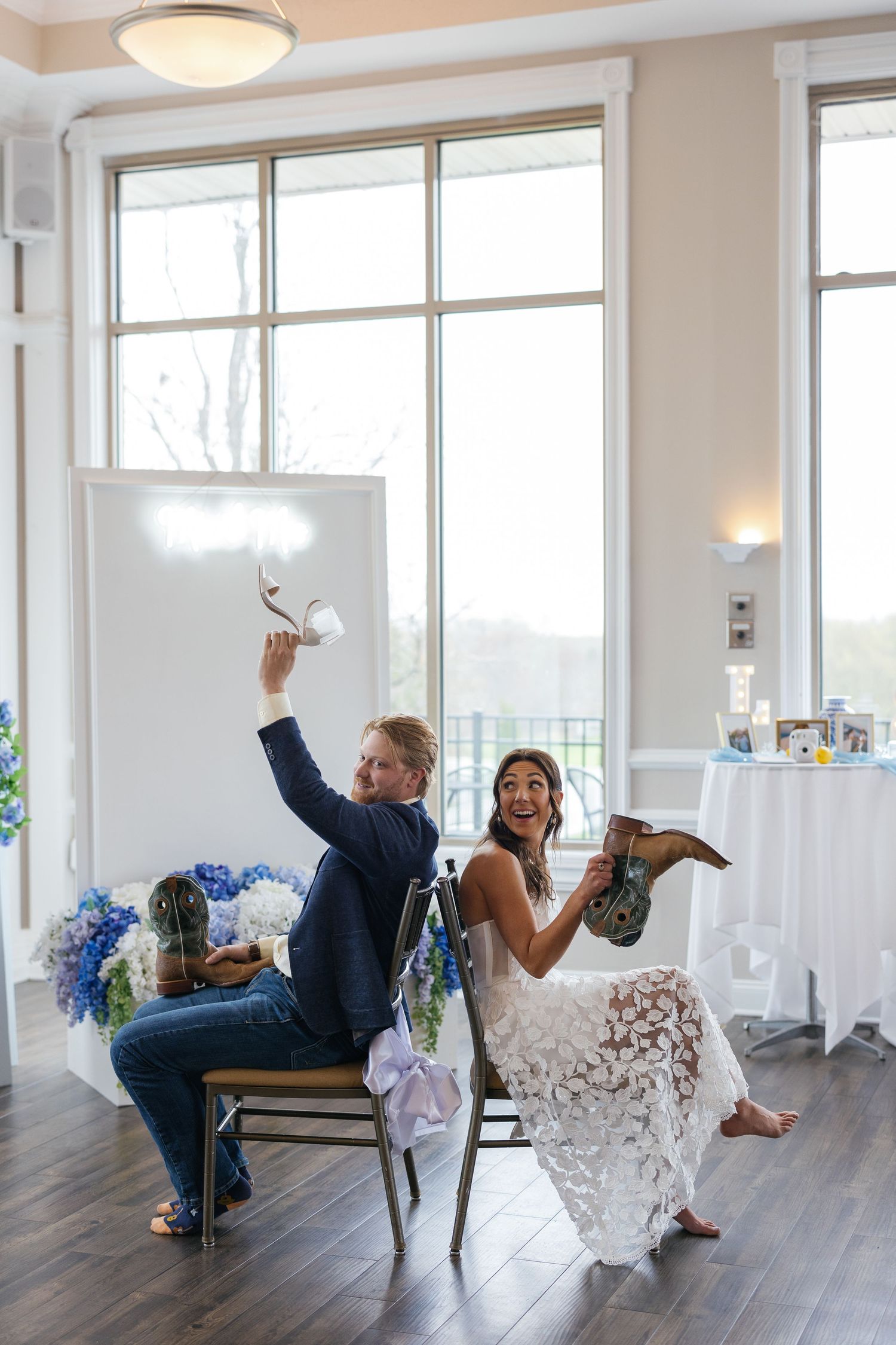 Playful wedding couple posing in chairs in a bright room with large windows.