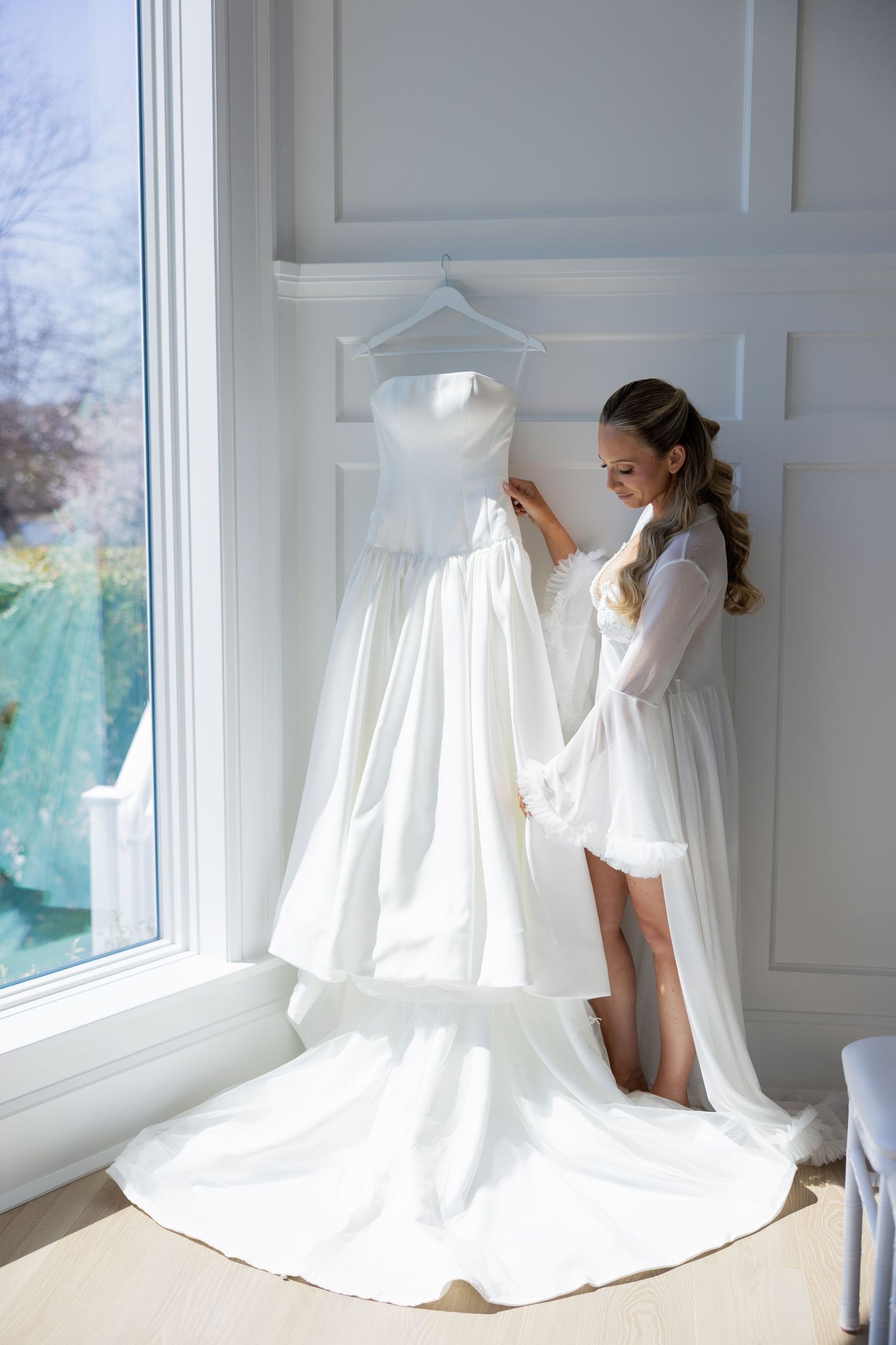 Bride in white robe admiring her elegant strapless wedding gown hanging by a bright window.