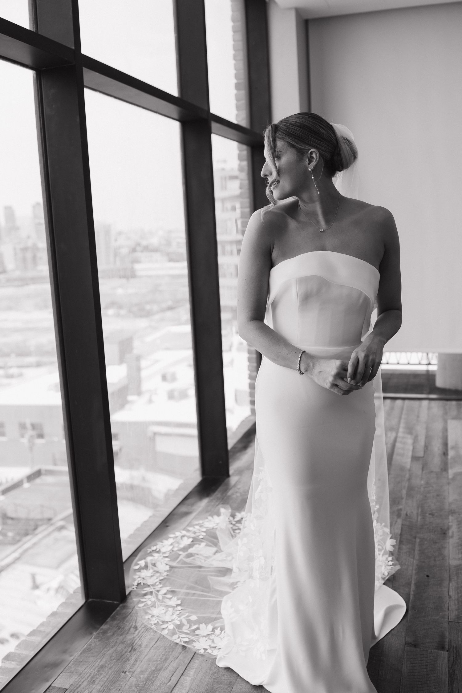 Elegant bride in a strapless white gown gazes out floor-to-ceiling windows overlooking a city skyline.