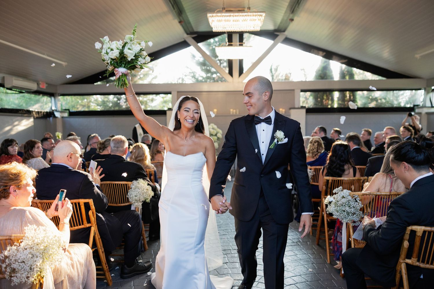 Bride and groom walk joyfully down the aisle after wedding ceremony, bride raising bouquet, guests seated in elegant venue.