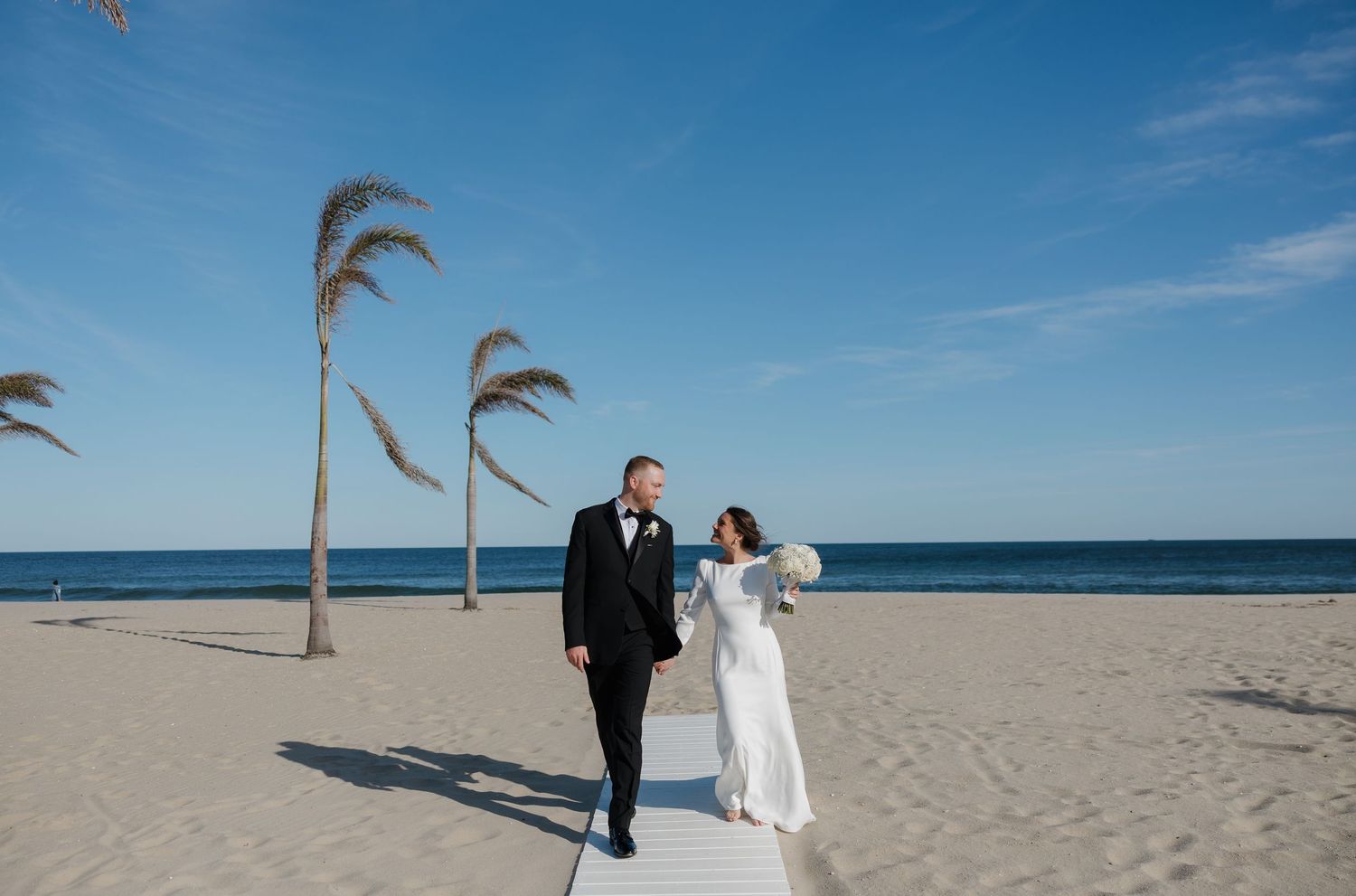 A couple walks along a sandy beach with palm trees and blue ocean waters on a sunny day.