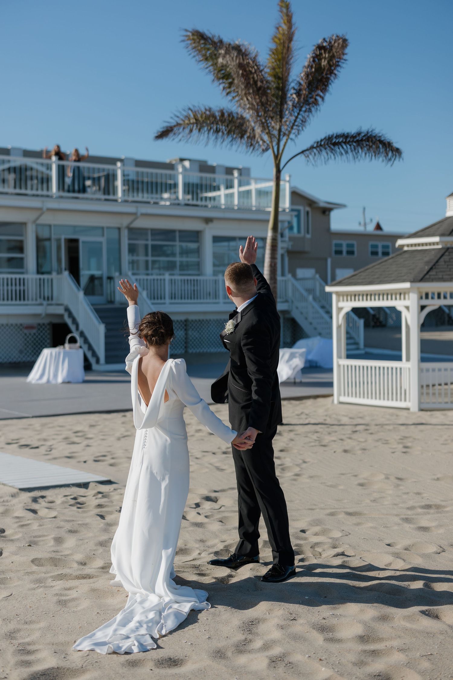 A couple shares a first look moment on a sunny beach with white buildings and palm trees in the background.