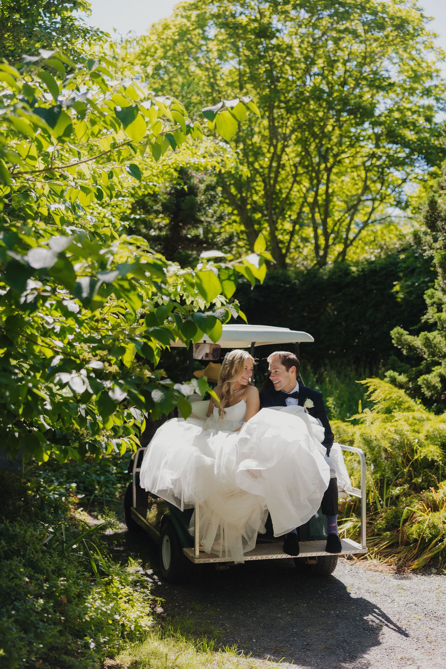 A couple in wedding attire sits together on a garden swing surrounded by lush green foliage on a sunny day.