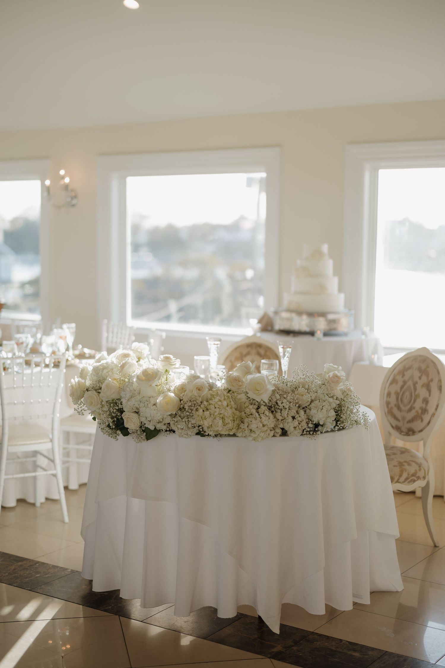 Elegant white wedding reception tables decorated with floral centerpieces and white linens in a bright sunlit room.