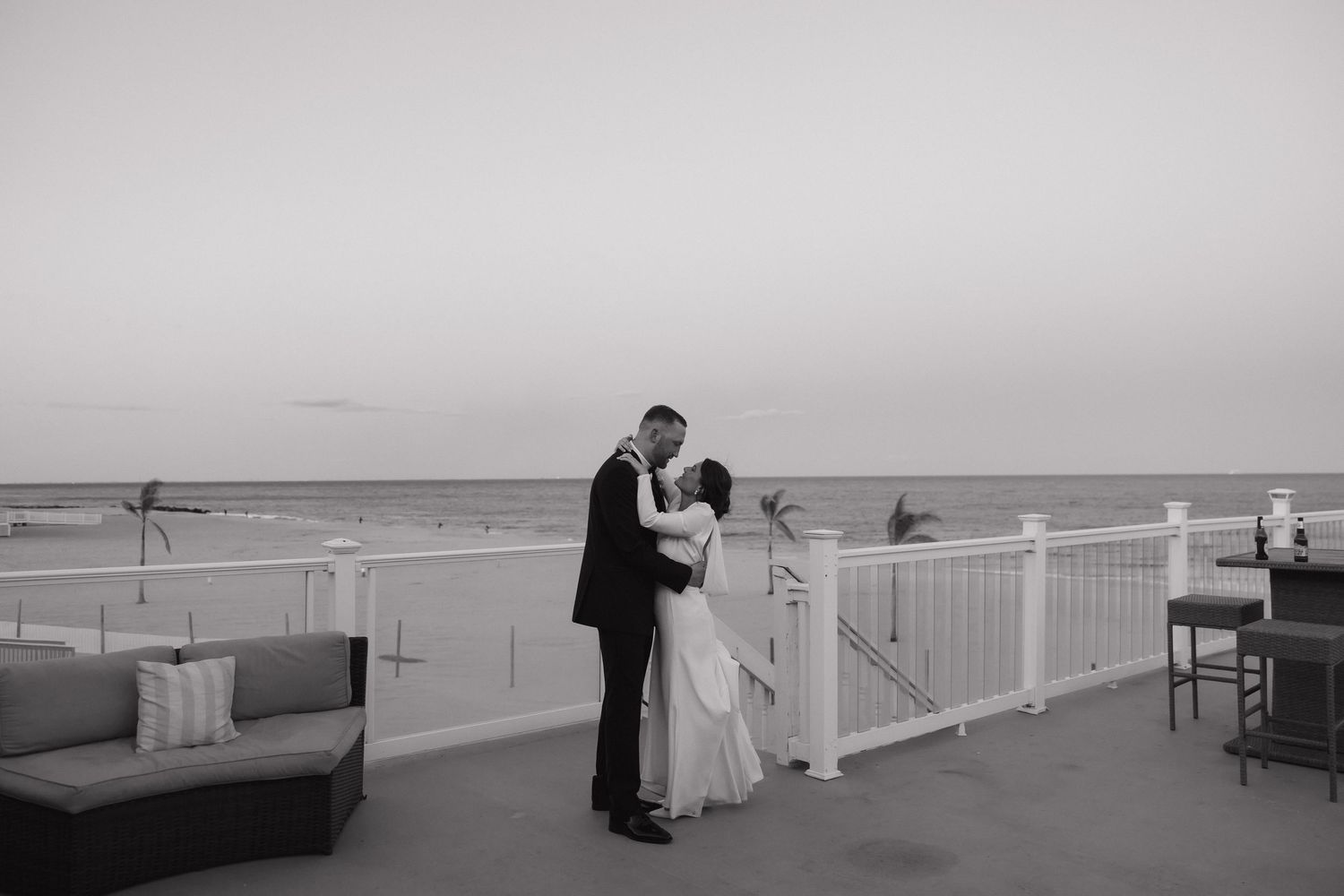A couple in formal wedding attire stands on a balcony overlooking a misty beach on their special day.