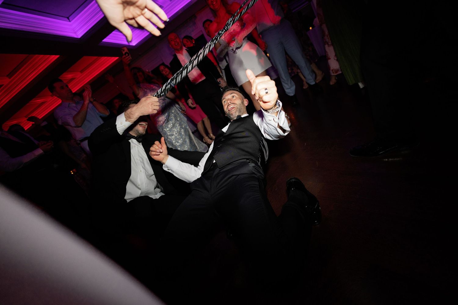 Groom in black tuxedo dancing energetically under limbo stick at lively wedding reception celebration.