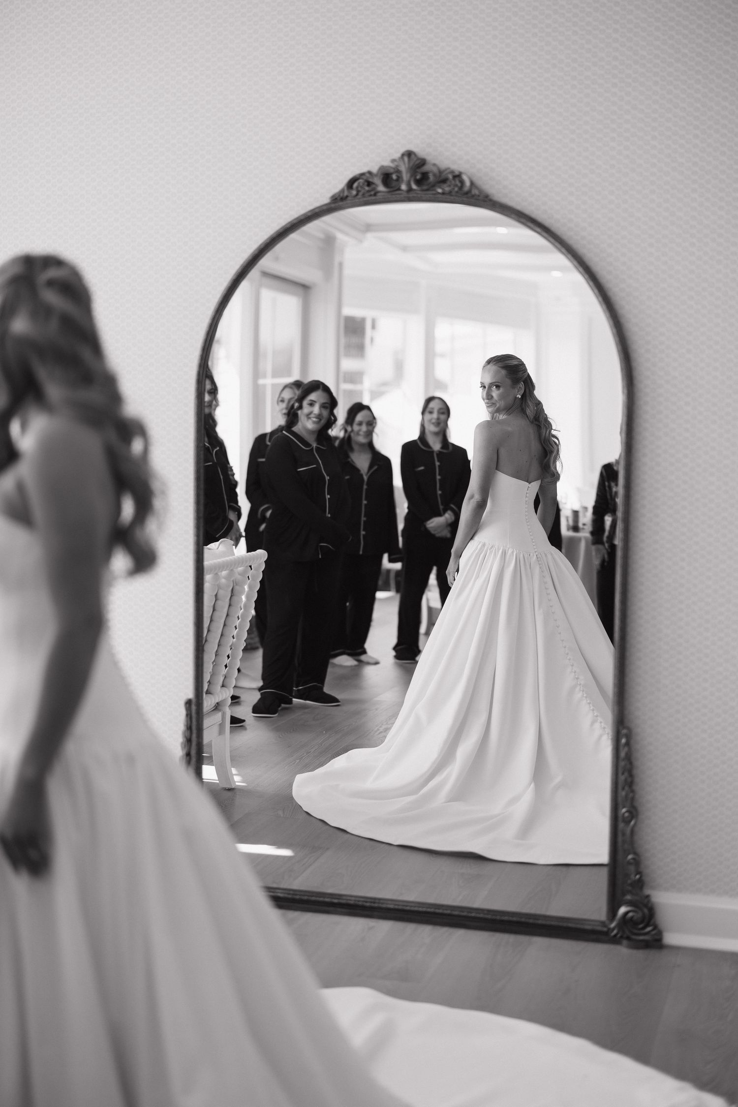 Black and white photo of bride in ballgown wedding dress reflected in ornate arched mirror with guests.