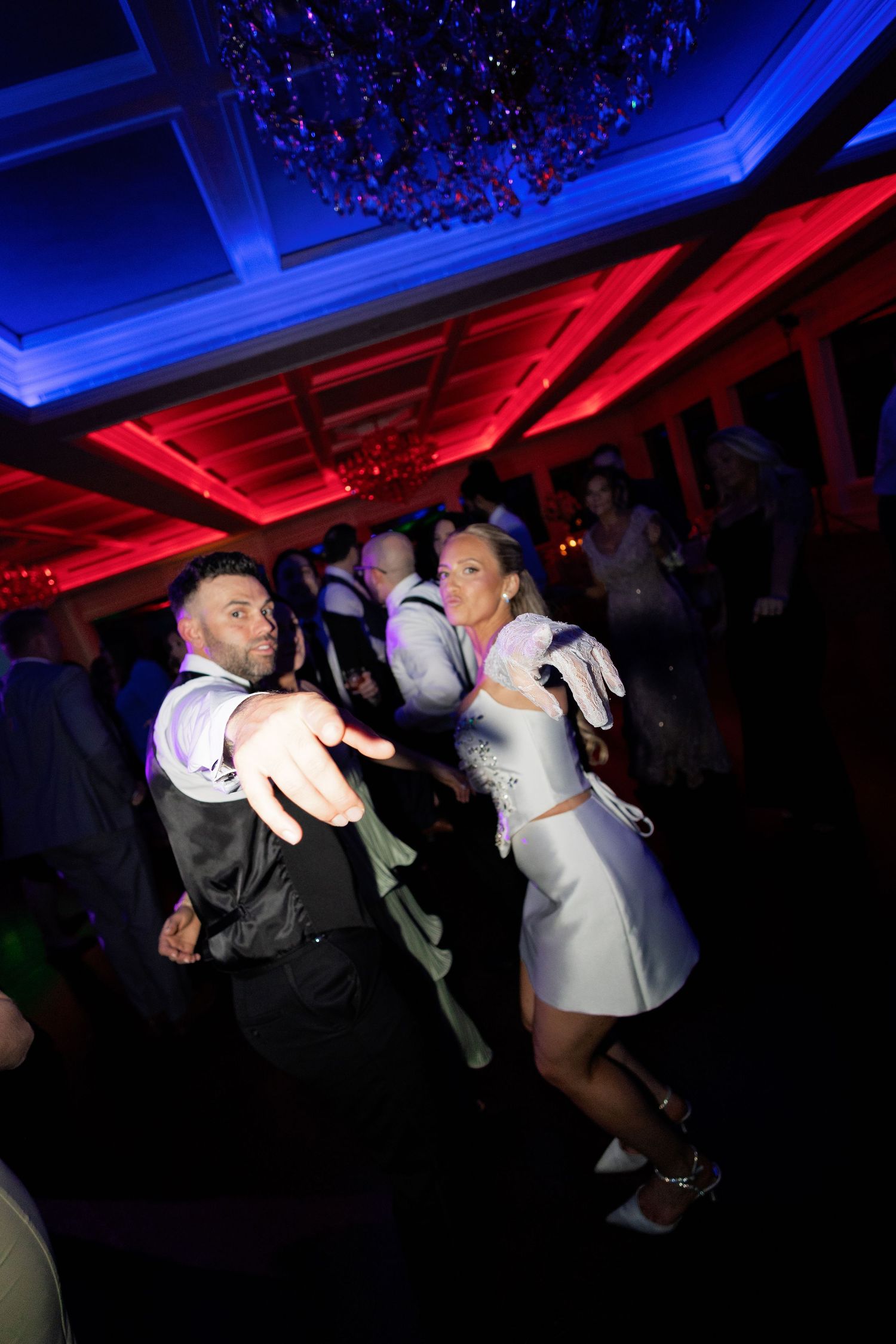 Bride and groom dance energetically with guests at their wedding reception under colorful blue and red lights.