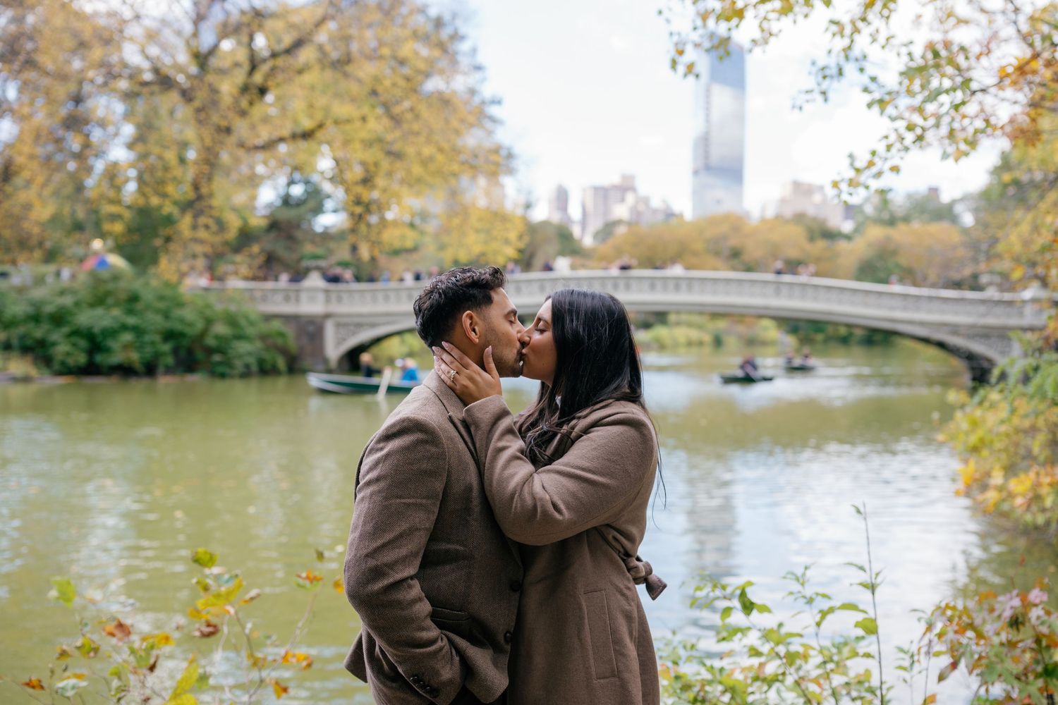 Couple sharing romantic moments by a scenic lake in Central Park during autumn with fall foliage in the background.