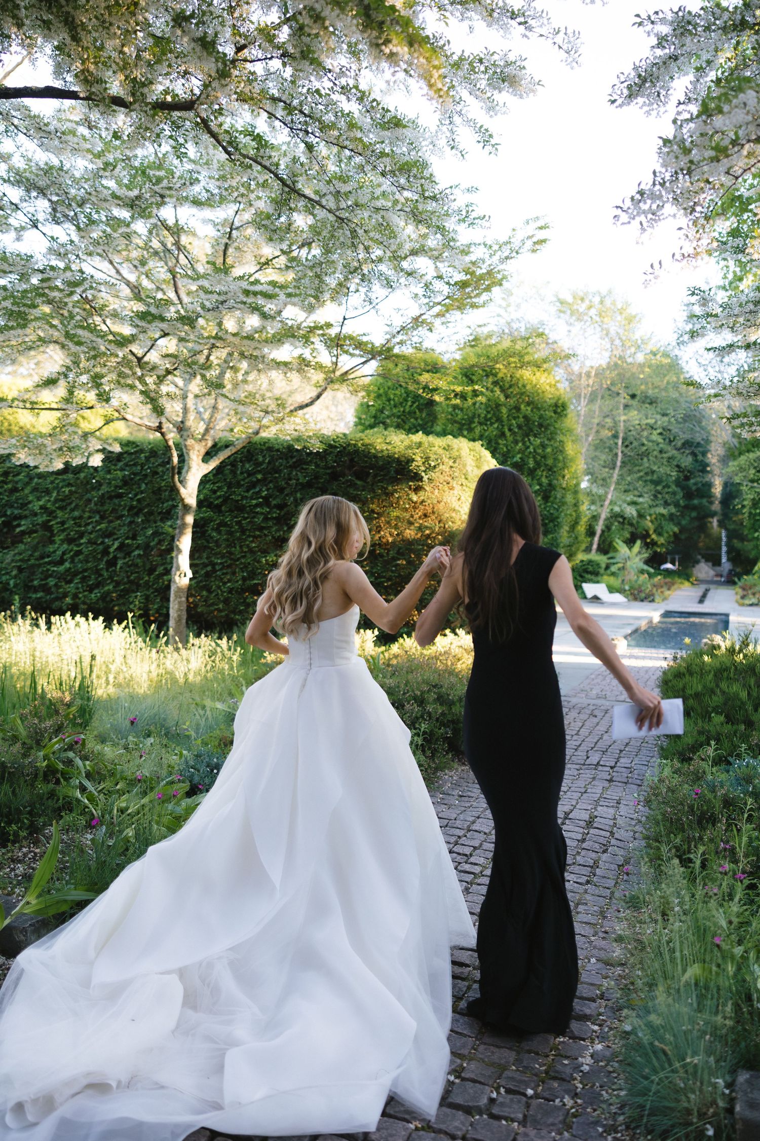 Wedding party walking along a garden path with flowing white dresses against green landscaping.