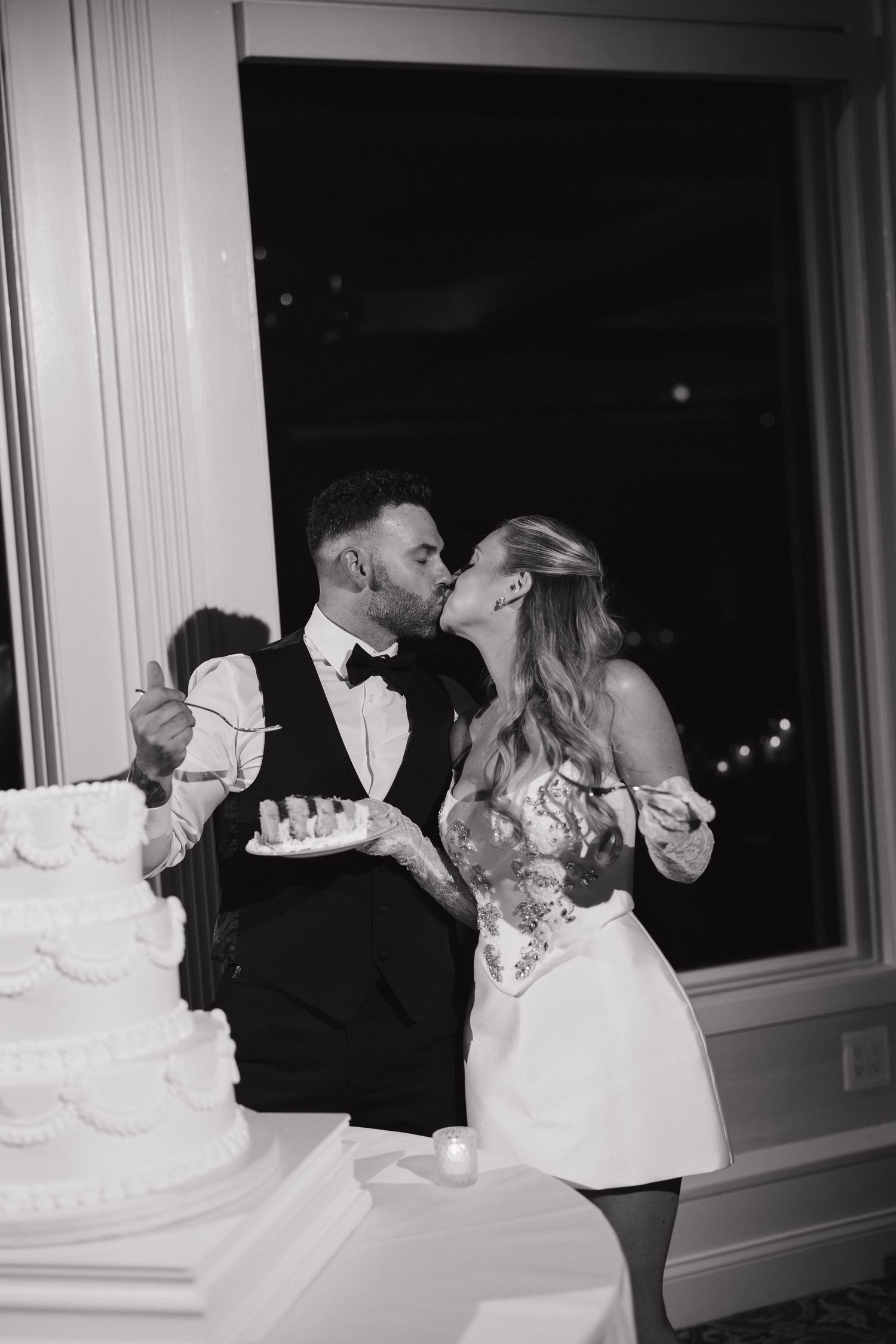 Black and white photo of bride and groom sharing a kiss while cutting their wedding cake at reception.